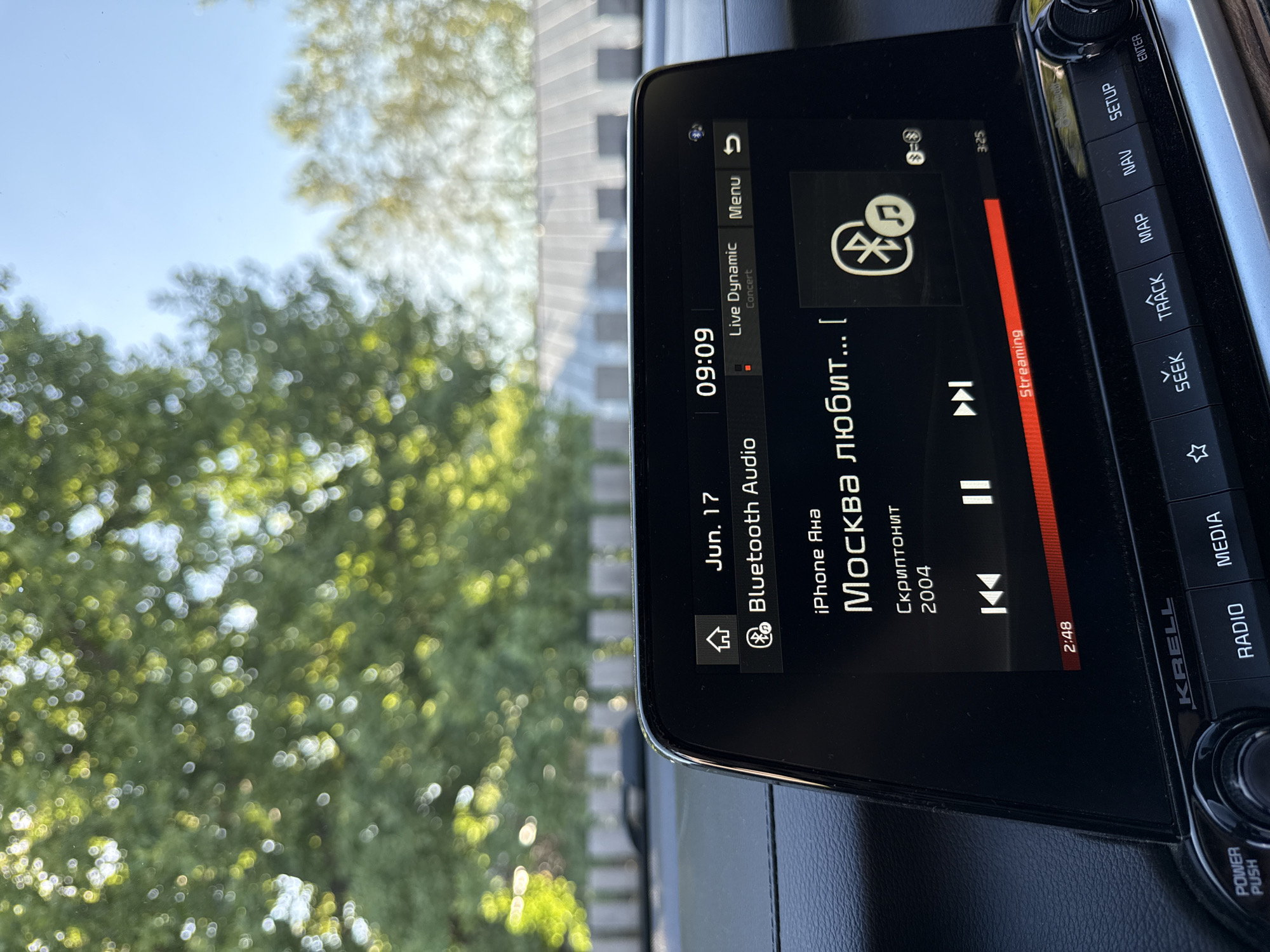 The image shows a car's infotainment system displaying a song titled "Москва любит..." by "Скриптонит" from the album "2004". The time is 09:09 on June 17th, and the audio is streaming via Bluetooth from an iPhone. The car is likely parked outdoors on a sunny day, as indicated by the dappled sunlight and foliage visible through the windshield. The surrounding environment suggests a residential or park area, possibly in the city of Chaikovsky, Russia, given the song's title referencing Moscow.