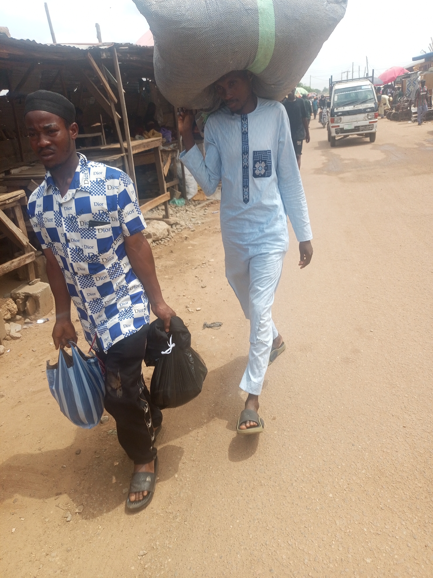 In Sabon Tasha, Nigeria, two men walk along a dirt road in a market. One man, dressed in a blue and white Dior-print shirt, black pants, and sandals, carries a blue striped bag and a black plastic bag. The second man, wearing a light blue, long-sleeved outfit and sandals, carries a large sack on his shoulders. In the background are market stalls and a small white truck. Other people are present in the background, some under umbrellas.