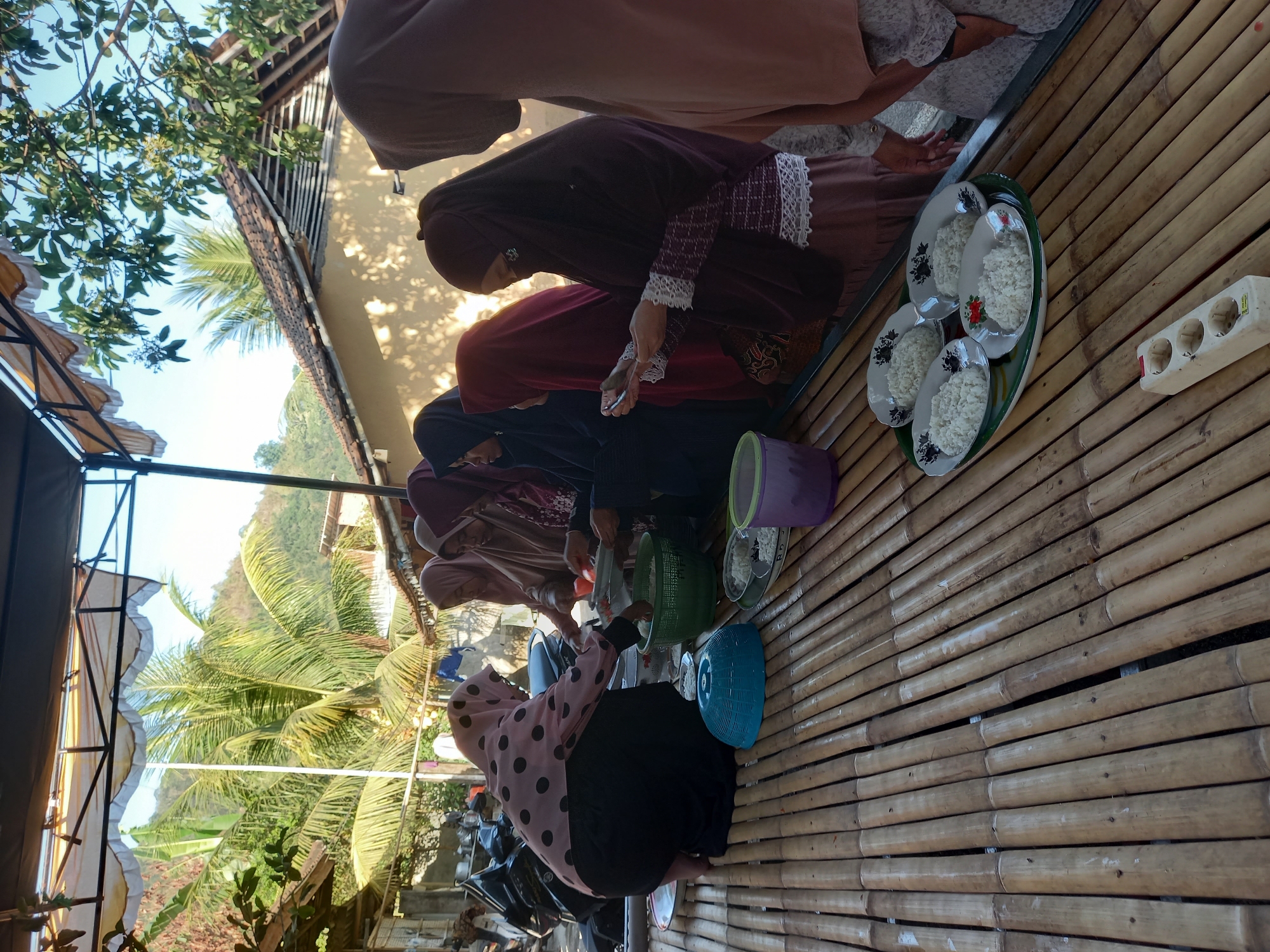 In Tempos, Indonesia, a group of women are gathered around a bamboo table, preparing to serve a meal. A woman in a pink hijab with black polka dots is kneeling and serving rice from a green bowl. Other women, all wearing hijabs in various colors including maroon, beige, and dark gray, are standing and observing. Some are holding plates and utensils. The table is laden with plates of white rice and a blue serving dish. In the background, a motorcycle is parked, and a hillside with trees and houses is visible. The scene appears to be an outdoor setting, possibly a residential area, with a clear blue sky above.