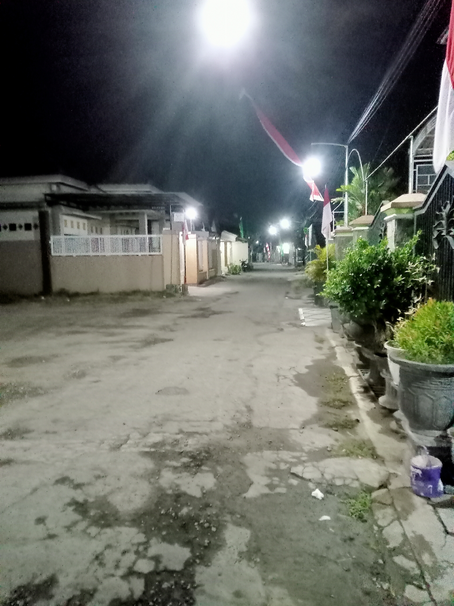 A night-time exterior shot of a street in Simo, Indonesia, likely a residential area, is presented. The scene is illuminated by a bright street light overhead, casting a strong glare and creating harsh shadows. The road surface appears to be asphalt or concrete, with visible patches of water and dirt, suggesting recent rain or a damp environment.
On the left side of the street, a single-story house with a light-colored facade and a white fence is visible. Several windows and an entryway are discernible. Further down the street, more houses and buildings line the road, some with lights on, contributing to the ambient illumination.
On the right side of the street, a black metal fence with decorative elements stands along the property line. Adjacent to the fence are several large potted plants containing lush green foliage. A few smaller potted plants are also present. In the foreground on the right, a purple bucket or container is situated on the ground near the edge of the road.
Several Indonesian flags, in red and white, are displayed along the street, attached to poles and seemingly fluttering in the breeze. These flags, along with the overall ambiance, suggest a festive or patriotic occasion. The sky is dark, typical of nighttime, with the bright street light being the dominant light source. The image has a grainy quality, indicative of low-light photography. No people are clearly visible in the scene.