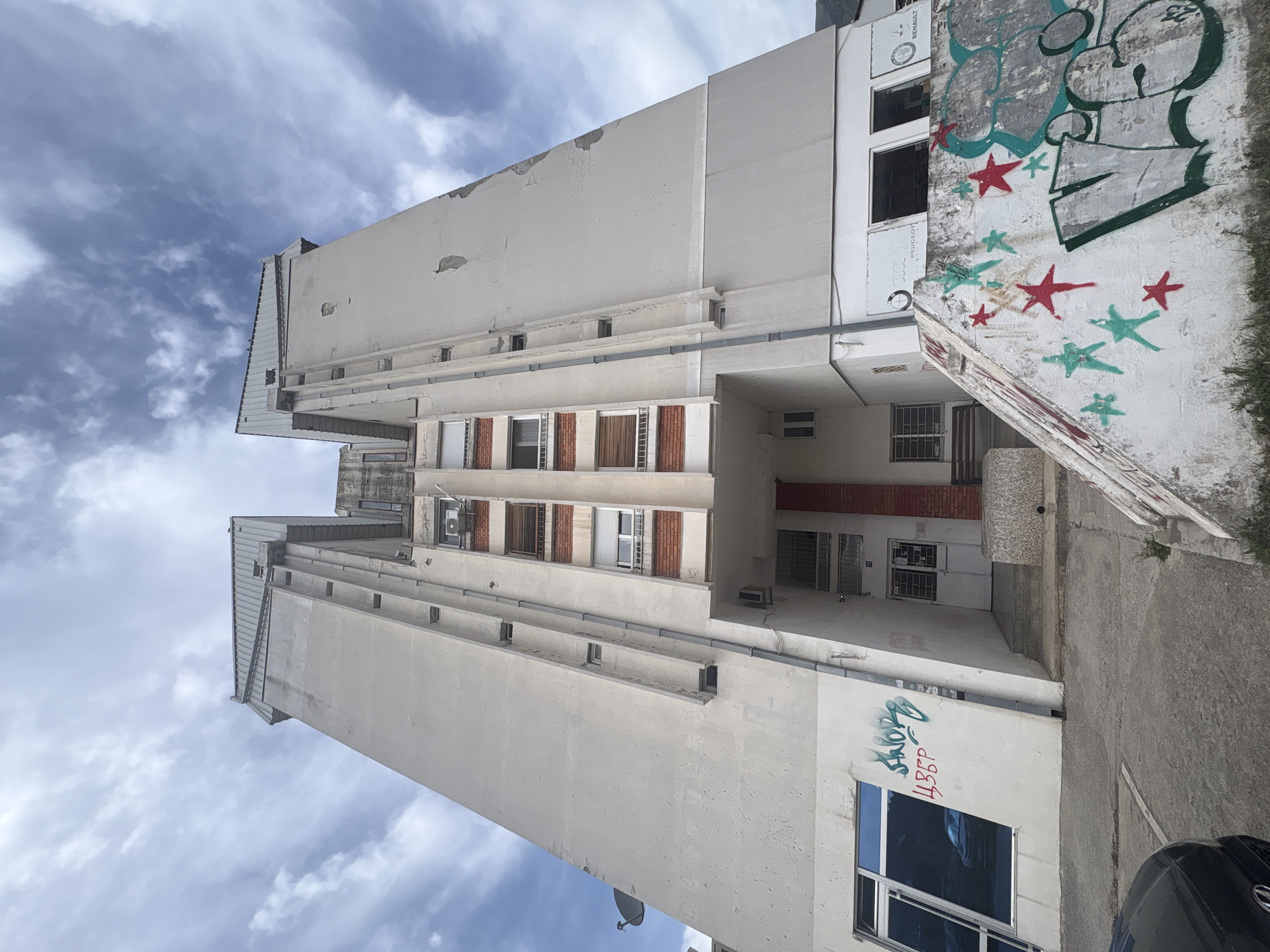 The image shows a tall, white, concrete apartment building with several stories, located in the city of Bar, Montenegro. The building appears to be in a state of disrepair, with patches of crumbling paint and visible wear on the exterior. There are windows of various sizes and styles along the facade, some with what appears to be brick detailing. A covered passageway is visible through the center of the building. Adjacent to the building is a concrete wall with graffiti, including stars, a cartoonish figure, and other tags. A portion of a black car is visible at the bottom right corner of the image, parked on what seems to be an asphalt or concrete surface. The sky above is partly cloudy. There are no people visible in the image.