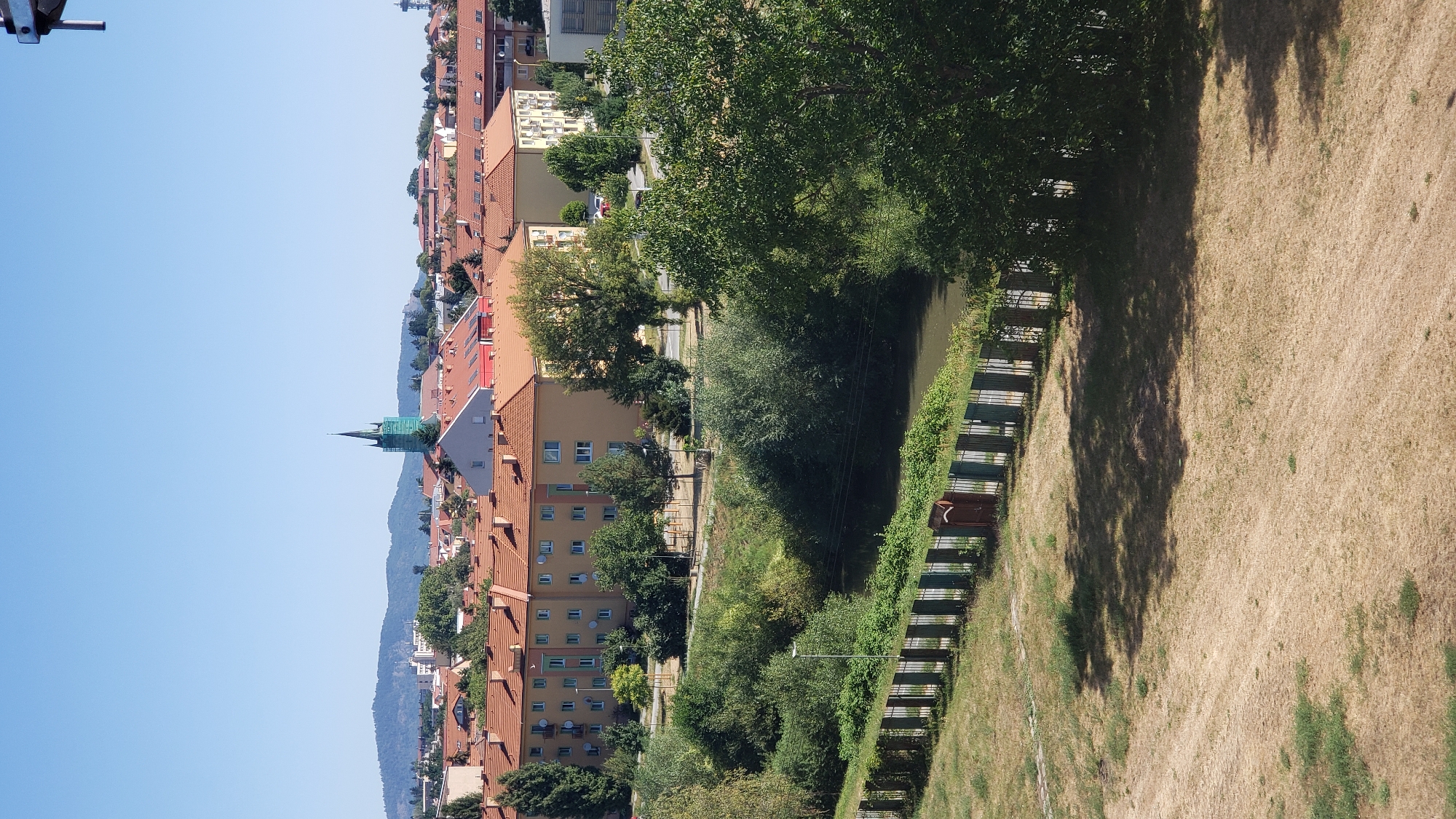 The image shows a panoramic view of a city under a clear blue sky. In the foreground, a dry, grassy field casts long shadows. A low fence separates the field from a river bank lined with lush green trees and bushes. Across the river, a row of apartment buildings with orange tiled roofs dominates the middle ground. A church steeple, painted green, rises above the buildings in the distance, flanked by more residential structures and a distant mountain range. The scene appears to be a typical urban landscape, possibly taken from a higher vantage point. The overall impression is one of a sunny day in a European city, with a blend of natural and built environments. Based on the church steeple and general architecture, this scene is likely Prešov, Slovakia.
