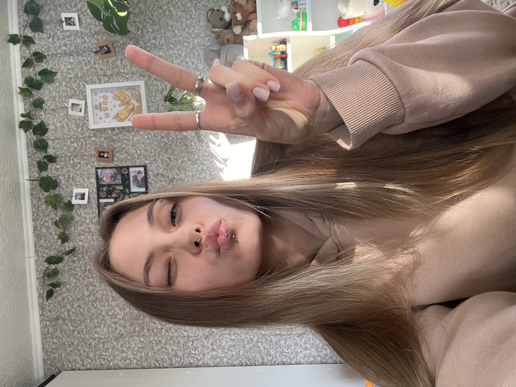 A young woman poses for a selfie indoors. She is fair-skinned with light brown hair and is wearing a beige sweatshirt. She winks one eye and puckers her lips. She has multiple piercings on her face: one through her eyebrow, one in her nostril, and two in her lower lip. Her hand is raised in a "peace sign" gesture, and silver rings are visible on her fingers. The background is a wallpapered wall adorned with artificial ivy and a string of photographs. A white shelf with toys is visible in the upper right. The image appears to have been taken in a residential setting. There is no verifiable information indicating the location is in Фоки, Russia.