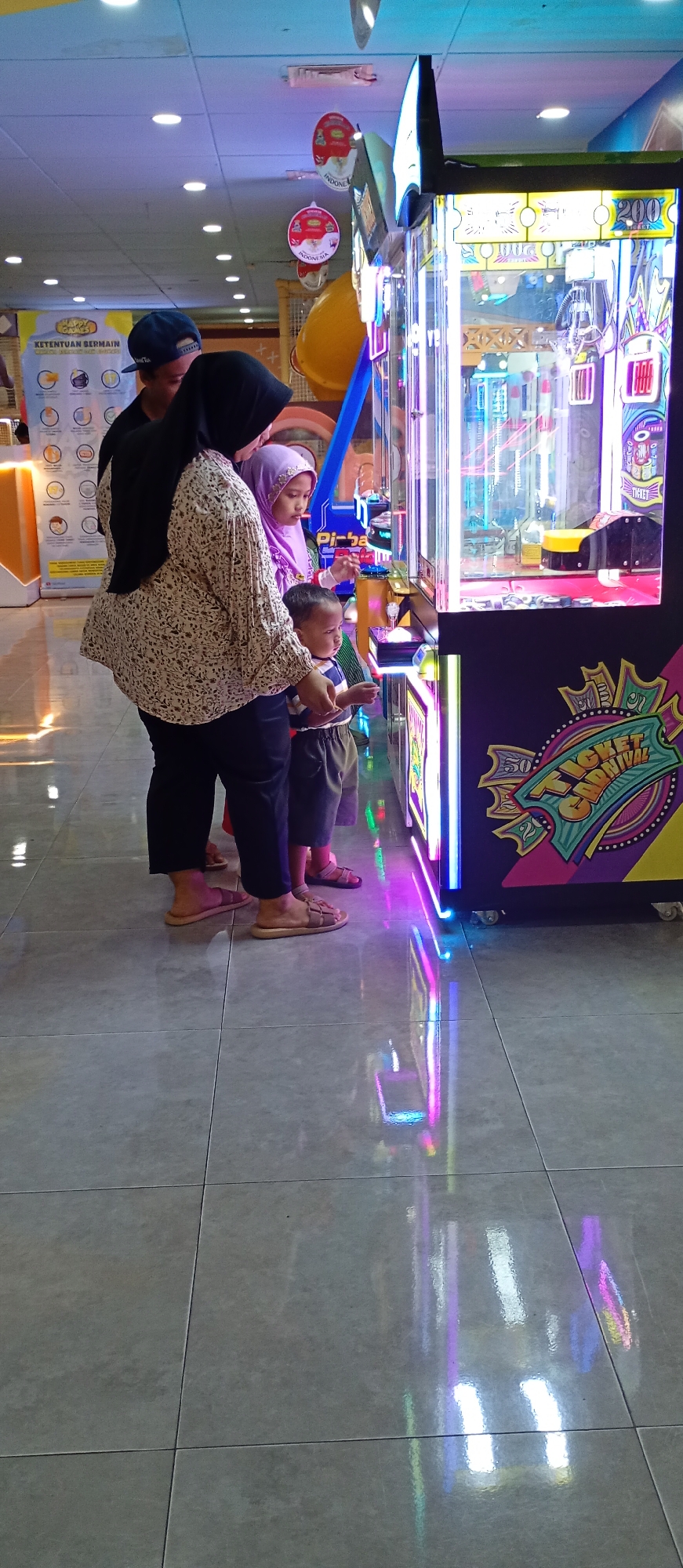 A family, likely composed of two parents and two children, stands before a brightly lit arcade game in a shopping mall or entertainment center in Probolinggo, Indonesia. The arcade game, with its vibrant neon lights and colorful graphics, features a claw machine mechanism. The mother, wearing a black hijab and a patterned blouse, stands on the left, her gaze directed towards the game. A young child, dressed in a striped shirt and shorts, stands between the mother and the game, appearing engaged with the machine. To the right of the child is a young girl, wearing a purple hijab and a light-colored top, also focused on the arcade game. A man, wearing a dark t-shirt and a blue baseball cap, is partially visible behind the mother, seemingly also observing the game. The floor is tiled and reflects the colorful lights of the arcade machine. In the background, a poster with text in Indonesian and circular graphics is visible. The overall scene suggests a casual outing at an entertainment venue.
