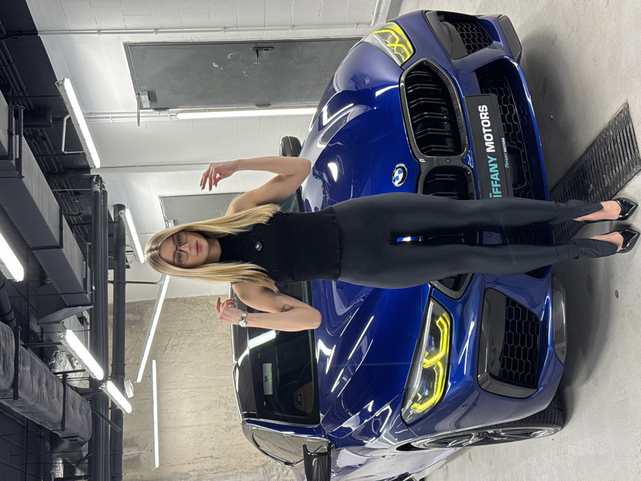 A blonde woman wearing a black shirt and leggings is posing on a blue BMW. She is standing with her legs extended and arms raised. The car is parked indoors with bright fluorescent lights overhead. The environment suggests a garage or dealership. The car has a "Tiffany Motors" sign on the front. The woman has glasses and is wearing a watch. The background is a mix of concrete walls and black pipes.