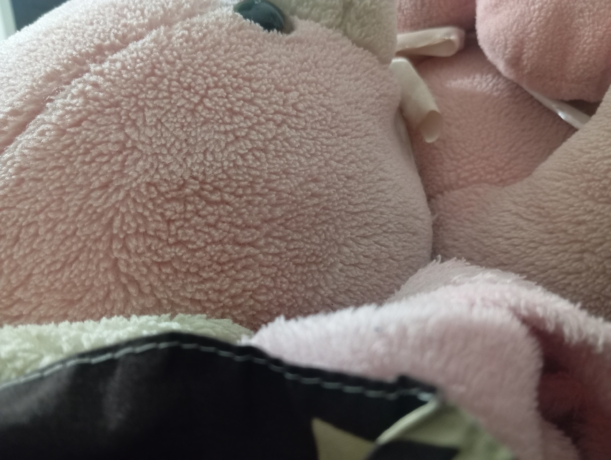 This is a close-up, high-angle shot of a pink, plush toy, likely a teddy bear. The toy is soft and fuzzy, with a textured surface. A seam runs diagonally across the toy's body. In the background, another plush toy or blanket is visible, also pink and fuzzy. Part of a dark object with white stitching is in the foreground, obscuring the bottom of the frame. There are no people visible in the image. The lighting is soft and diffused, suggesting an indoor environment. The specific location in the city of Rejosari, Indonesia, is not discernible from the visual information provided.