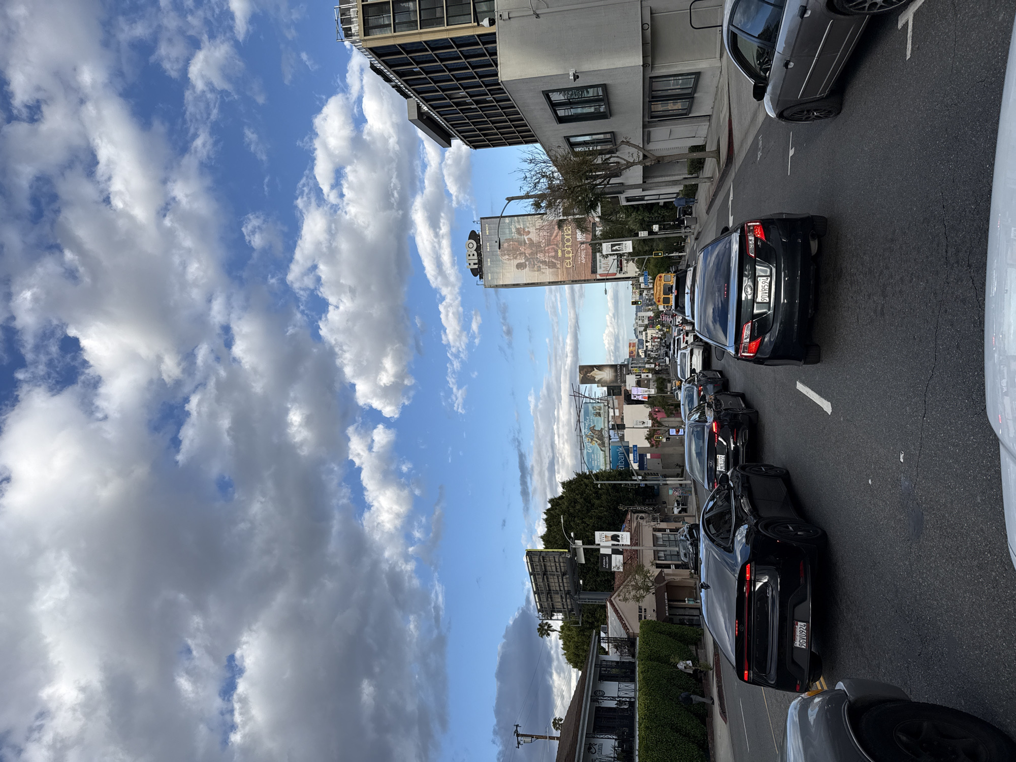 The photograph, taken at street level in West Hollywood, United States of America, depicts a congested street filled with stationary cars during daylight. The sky, visible above the buildings and billboards, shows a mix of blue sky and voluminous white clouds. Several large billboards advertise various media products, including a billboard for HBO's "Euphoria." Buildings of varying heights line the street. The cars, mostly black, gray, and white, are tightly packed, indicating heavy traffic. The environment suggests an urban setting, likely a commercial or entertainment district. No people are clearly visible, though some are partially obscured.
