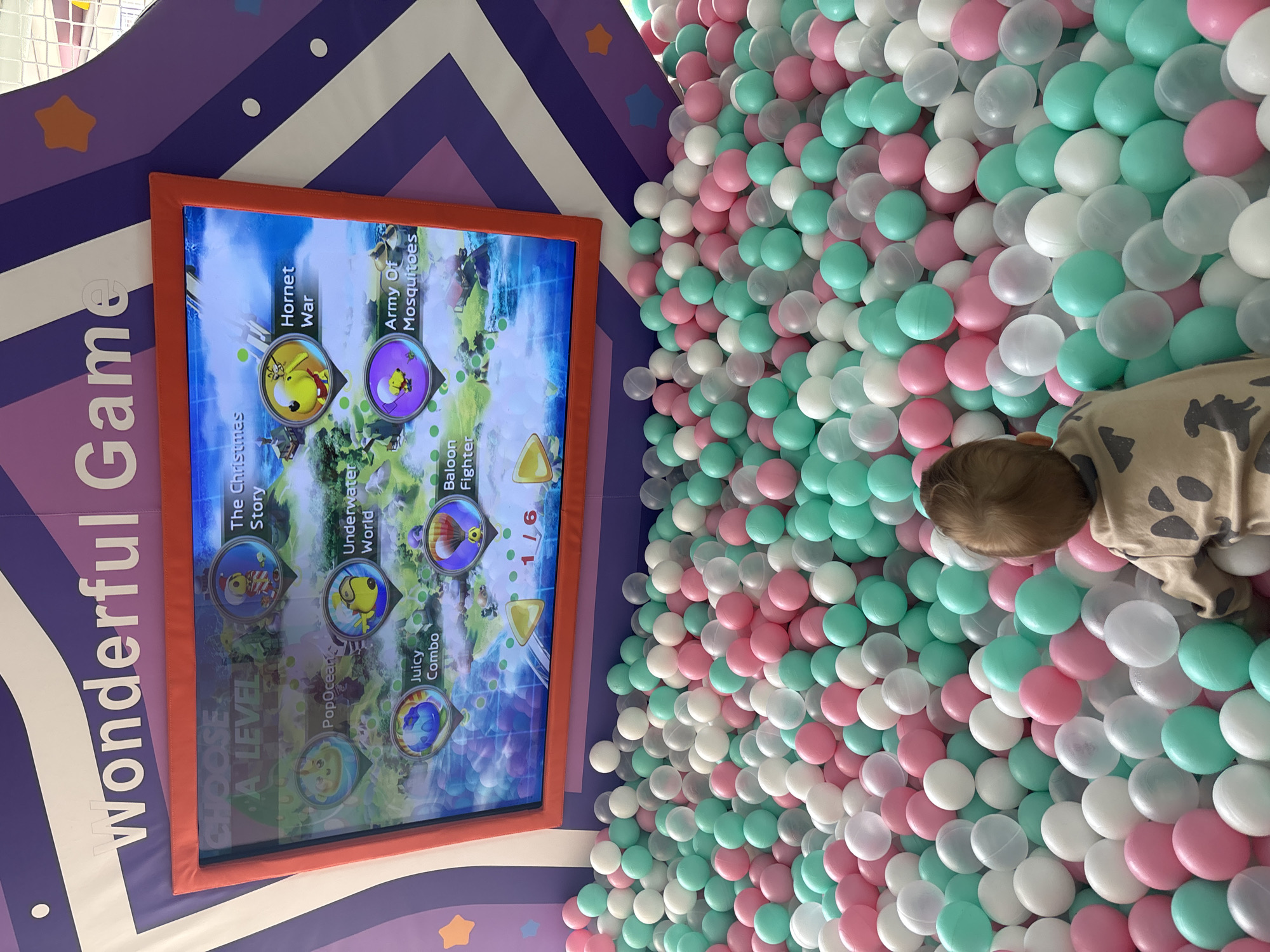 The image depicts a child partially visible in a ball pit filled with pastel pink, white, and light green balls. Adjacent to the pit is a digital screen displaying a game menu titled "Wonderful Game" with options like "The Christmas Story," "PopOcean," "Underwater World," "Juicy Combo," "Baloon Fighter," "Hornet War," and "Army Of Mosquitoes." The screen is surrounded by a purple, white, and orange patterned border. The location is likely an indoor play area, and the image was taken in Perm, Russia.