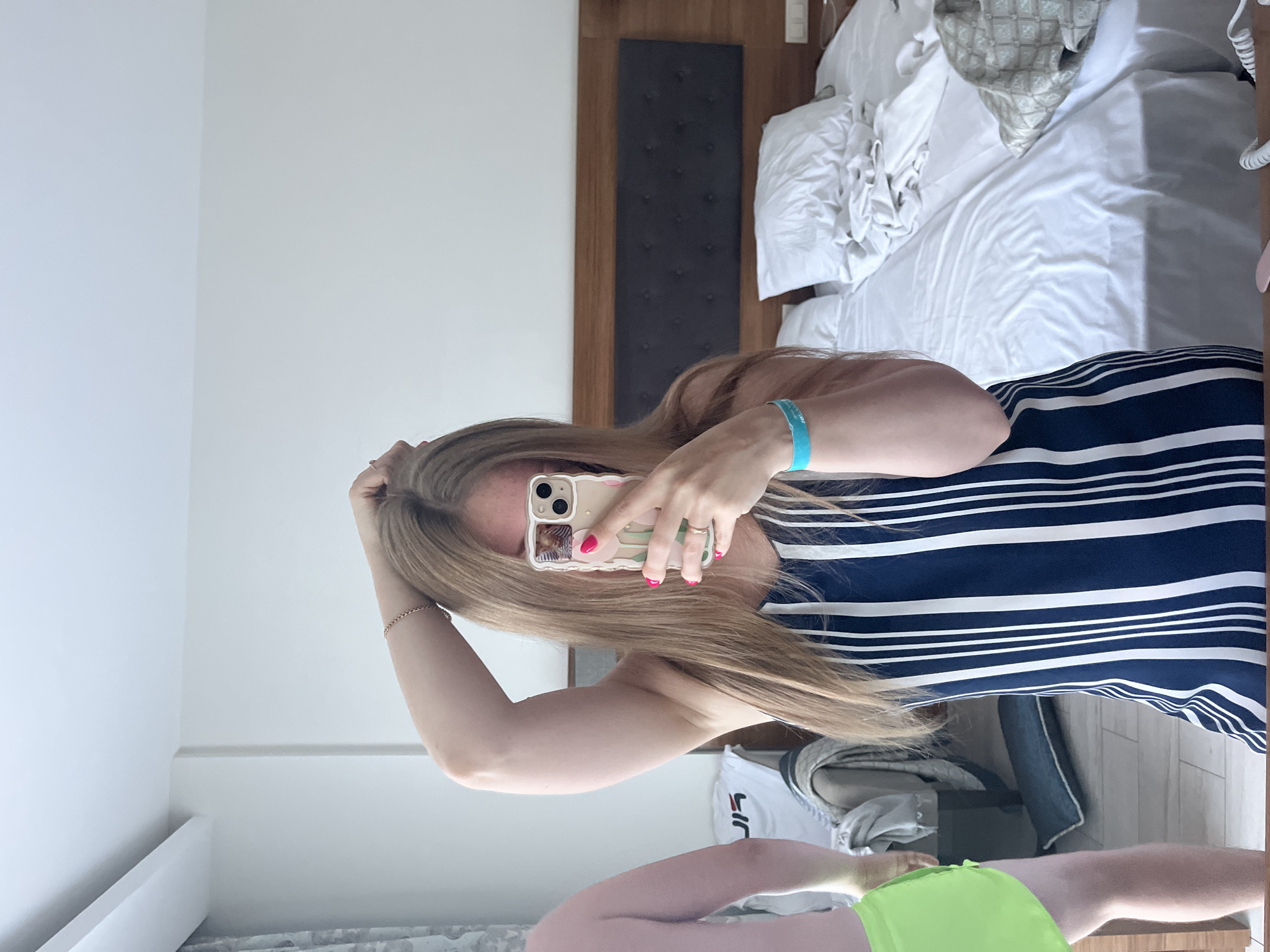 A blonde woman, wearing a blue and white striped dress, neon green shorts, and a turquoise wristband, poses for a selfie in a hotel room in Alanya, Turkey. She holds her phone in her right hand, her left arm raised above her head. A man, partially visible in the reflection, stands to her left, wearing neon green shorts. The room features a large bed with white linens, a wooden headboard, and a dark upholstered accent. A chair with clothing draped over it is visible in the background, along with a curtain and part of a window.