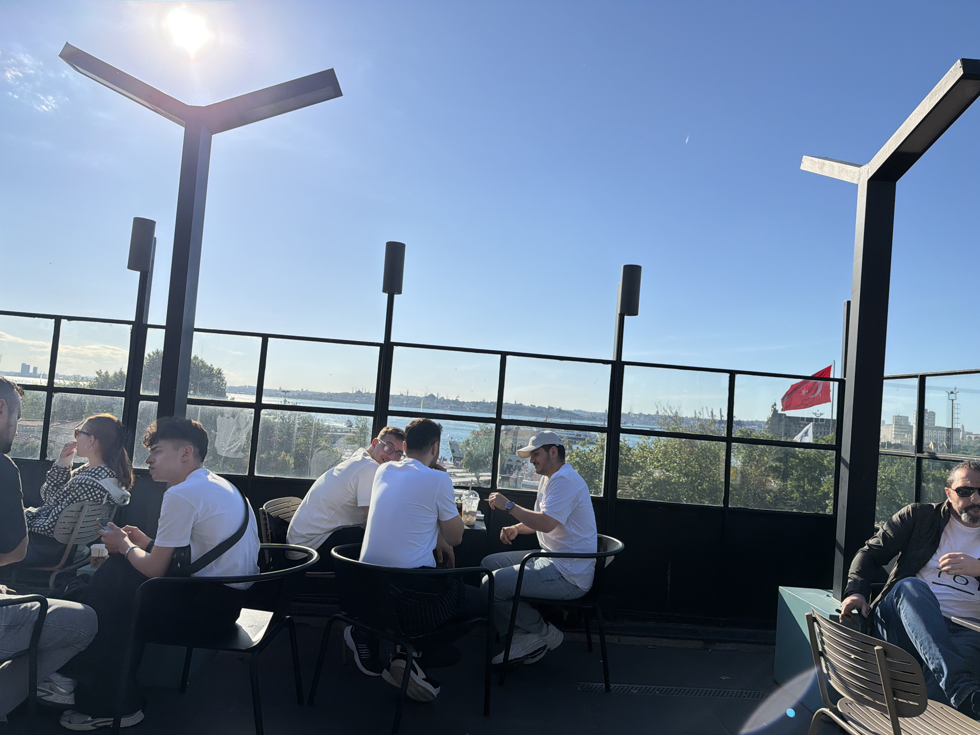The image shows a group of people sitting on a rooftop terrace in Kadıköy, Turkey. The terrace has a black metal railing with glass panels, offering a view of the cityscape and the sea. There are several men wearing white t-shirts sitting at a table and others are sitting around. A Turkish flag is visible in the background. The sky is clear and blue with a bright sun. Some architectural structures are also visible on the terrace.