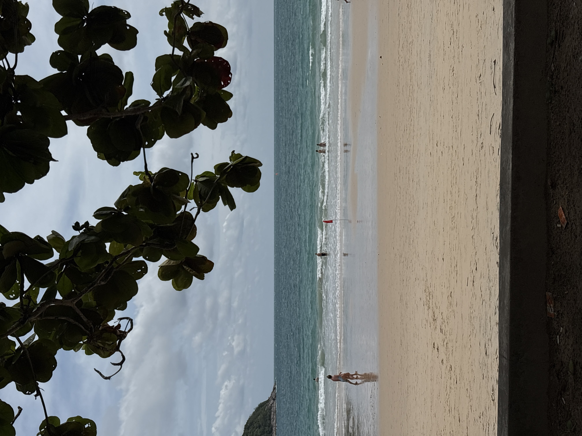 The image depicts a beach scene in Rawai, Thailand. The shot is taken from a slightly elevated position, partially obstructed by foliage in the left foreground. The beach is sandy, with gentle waves rolling in. Several people are visible at different points along the shoreline, some wading in the water. A red flag is planted in the sand near the middle of the frame. The sky is overcast, with a grayish hue. A landmass covered in vegetation is discernible in the distance. The right edge of the frame shows a dark, textured vertical element, potentially a wall or embankment.