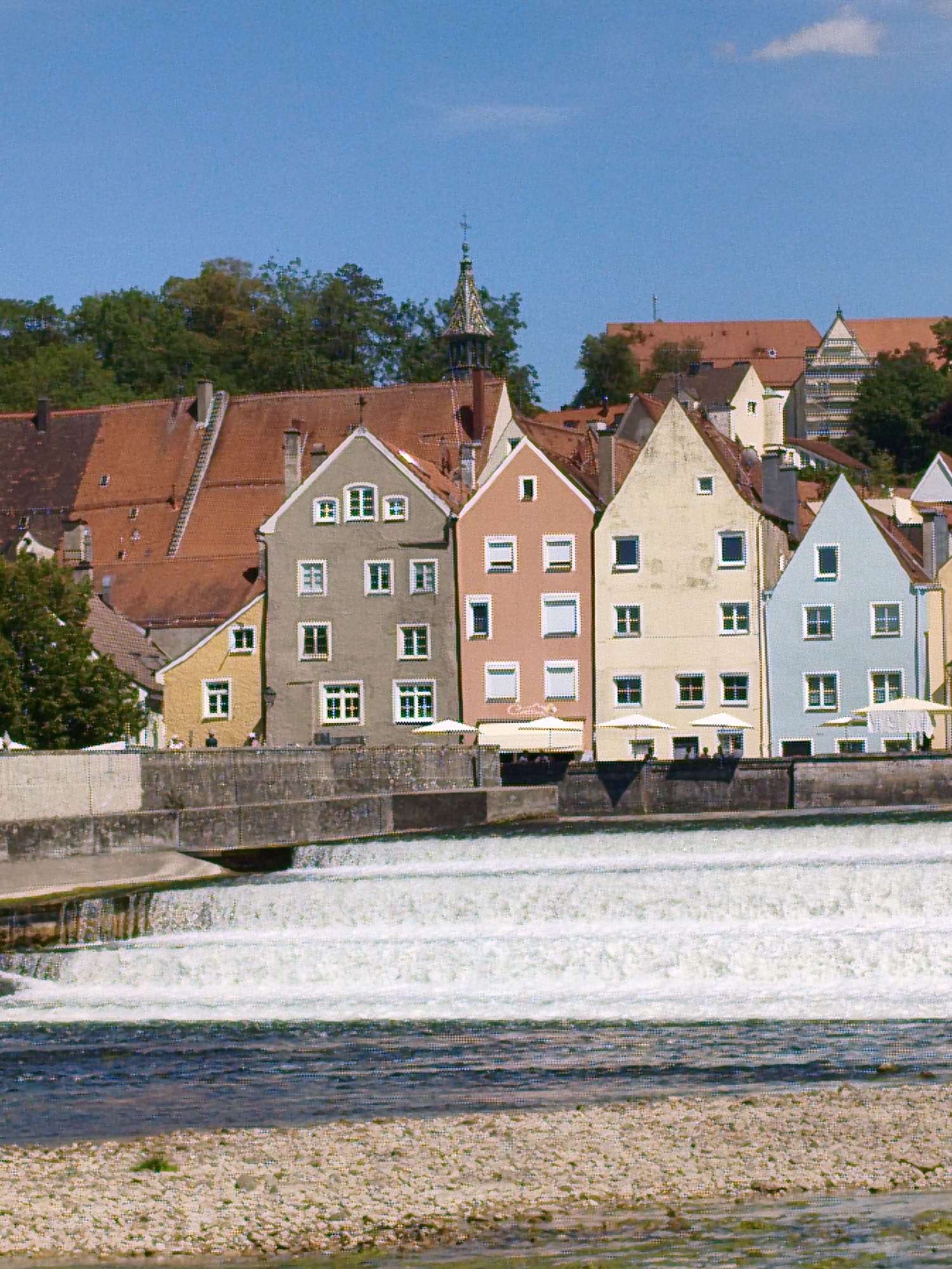 The image captures a scenic view of the Lech River in Landsberg am Lech, Germany. In the foreground, the river flows over a man-made cascade, creating a white, frothy surface. The riverbed is visible in the bottom portion of the image, composed of various sized pebbles. A stone embankment lines the river to the left and right.
Above the embankment, a row of historic, colorful buildings stands prominently. These buildings feature multi-story facades, pitched roofs with red tiles, and numerous windows. The buildings are painted in various shades of grey, yellow, coral, beige, and light blue. Some of the buildings have awnings or umbrellas extending from their fronts, suggesting outdoor seating for businesses.
Behind the row of buildings, a dense green foliage of trees forms a backdrop, with a glimpse of a church spire with a decorative roof peaking through the trees. The sky above is a clear, bright blue with a few wispy white clouds. The overall scene depicts a charming European town situated alongside a flowing river, characterized by its architecture and natural landscape.