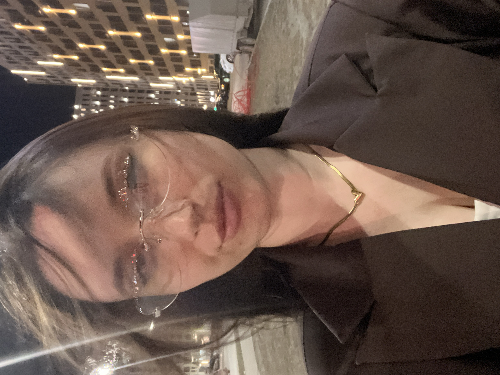 A medium shot depicts a woman with glasses, wearing a brown jacket and a gold necklace, likely taken at night in Tyumen, Russia. The background shows a modern building with illuminated windows and streetlights. The woman's face is partially visible, with a focus on her glasses and facial features. The jacket suggests it may be a chilly evening. The overall impression is that the photo was taken casually, possibly as a selfie or by someone nearby.