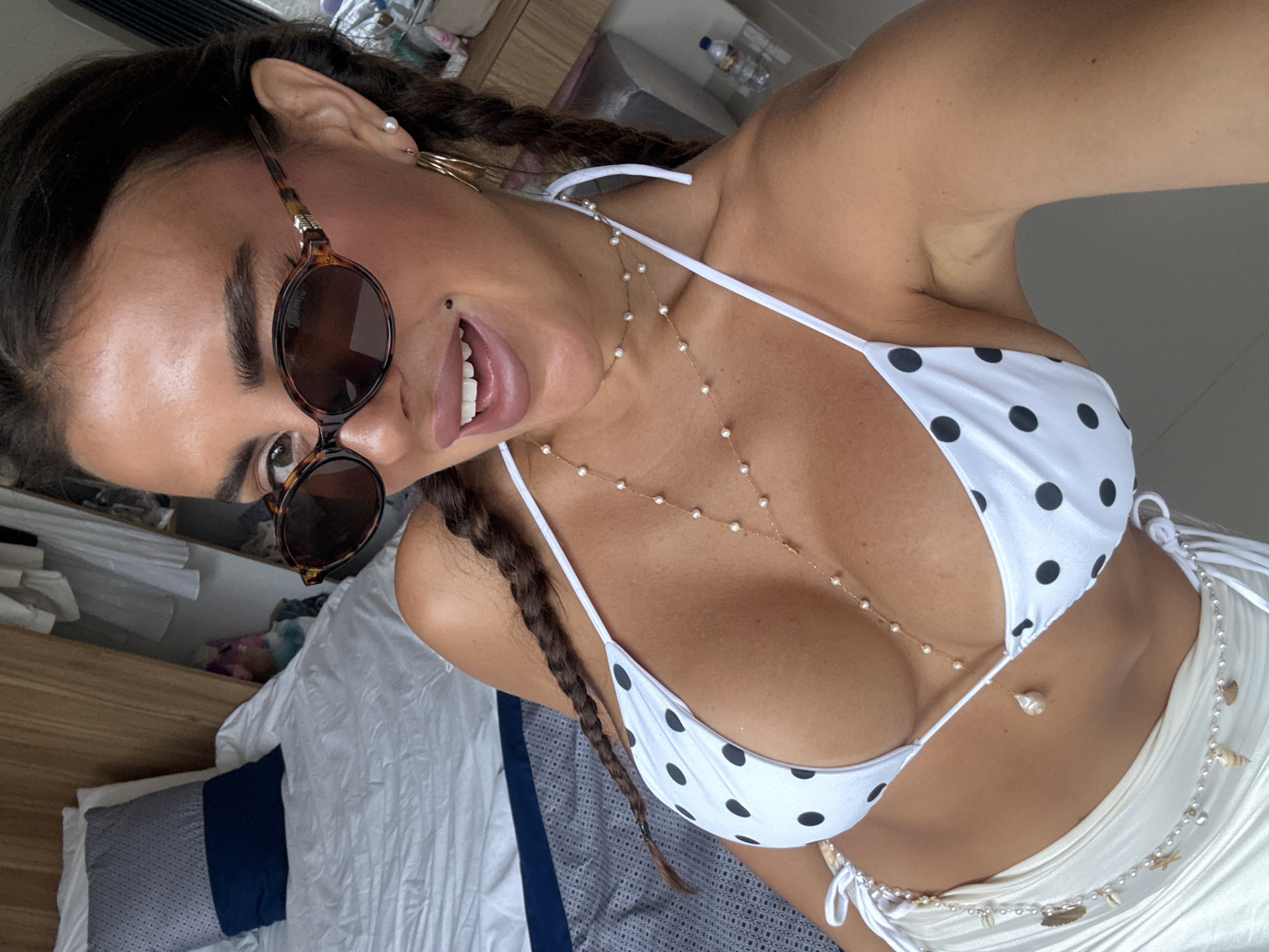 A selfie angled from above shows a fair-skinned woman with dark hair styled in braids wearing a white bikini with black polka dots and round, brown sunglasses. She is also wearing jewelry including a necklace with small pearls and a body chain with seashells. The background includes a bed with blue and grey bedding and a shelf with miscellaneous items. Location information is not discernible from the image.