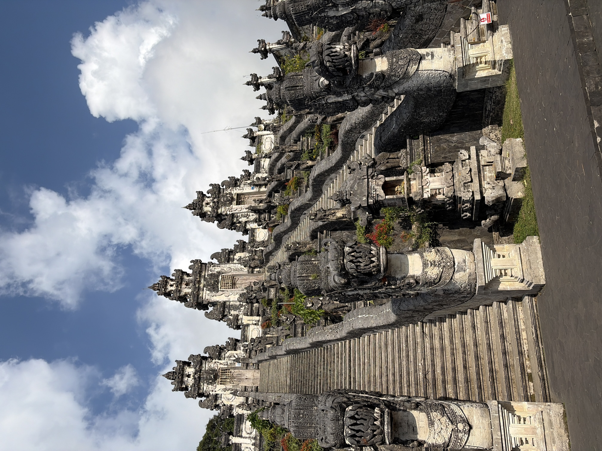 The image depicts the grand entrance to a temple complex, characterized by a long, wide stone staircase flanked by ornate dragon sculptures. The stairs ascend towards intricately designed temple structures with tiered roofs and decorative carvings. The architecture suggests a Balinese style, with details like narrow doors and statuary adorning the walls. Lush green foliage and small bursts of red flowers are visible along the sides of the stairs, adding color to the predominantly stone and grey landscape. The sky above is a vibrant blue, dotted with scattered white clouds, indicating fair weather. The foreground shows a paved pathway leading to the base of the stairs. The overall scene evokes a sense of ancient grandeur and spiritual significance. The location is identified as Abang, Indonesia, suggesting this is likely a significant religious site within that region. There are no people visible in the image.
