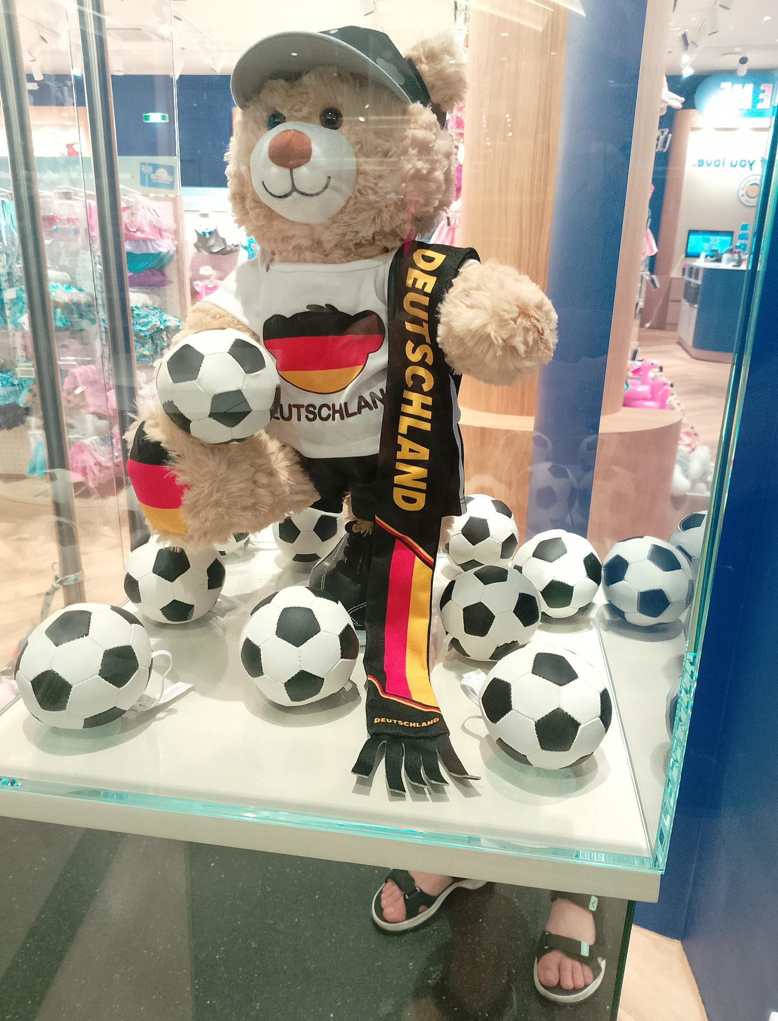 A teddy bear wearing a cap and a shirt with the German flag and "Deutschland" emblazoned on it is displayed in a store window, surrounded by several soccer balls. The teddy bear is holding a soccer ball and has a scarf with the German flag colors. The display is on a white shelf with a blue light strip. Feet wearing sandals are visible at the bottom of the image, suggesting a shopper is looking at the display. The background shows shelves with clothing and other merchandise.