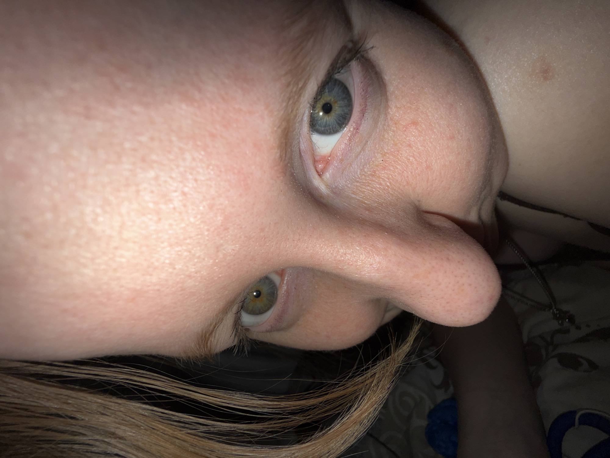 A close-up, slightly off-center image of a person's face, angled downwards. The subject has light skin, with visible pores and some freckles. The left eye (from the viewer's perspective) is blue, while the right eye appears greenish-hazel. Light brown hair is visible in the lower left corner of the image. A necklace is seen near the right side of the image. The background is dark and out of focus, but a bed sheet with a pattern is visible. The location is not discernable from the image and cannot be determined to be Perm, Russia.