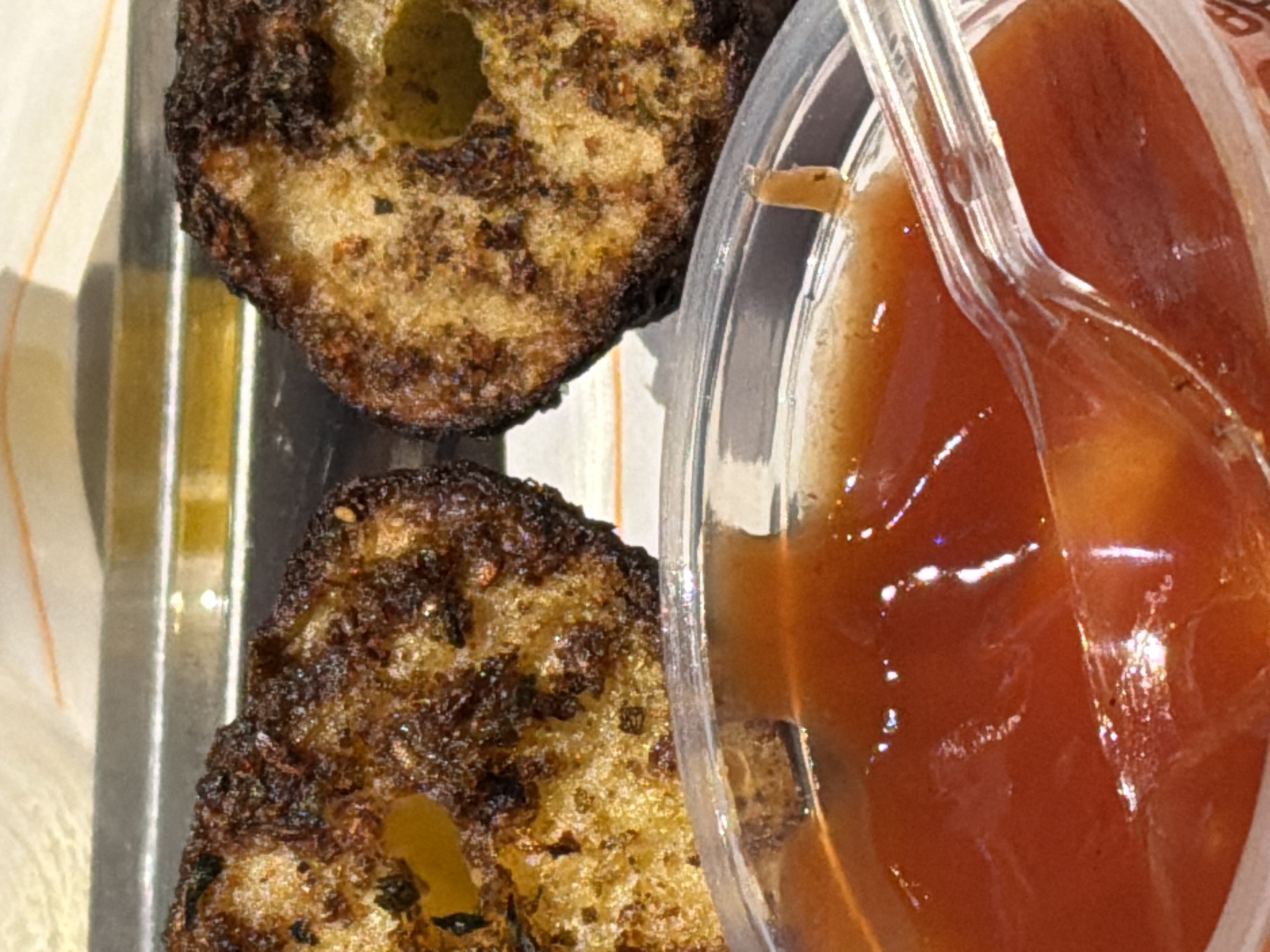 A close-up, slightly overhead shot captures two pieces of fried or baked food next to a container of sauce, set on a metallic tray. The food items, possibly fritters or patties, feature a dark, heavily browned and textured crust, suggesting deep frying or intense baking. Their interiors appear lighter, golden-brown, and somewhat porous. Next to them, a small, clear plastic cup holds a viscous, reddish-brown sauce, which could be ketchup, barbecue sauce, or a similar condiment. A translucent plastic spoon is partially submerged in the sauce. The metallic tray reflects some light, and a subtle pattern on what seems to be a paper liner or napkin is visible underneath the food. The background is softly out of focus, indicating a shallow depth of field. No people are visible, and no discernible text appears in the image. The scene suggests a casual meal, possibly in a restaurant or take-away setting in Yerevan, Armenia.