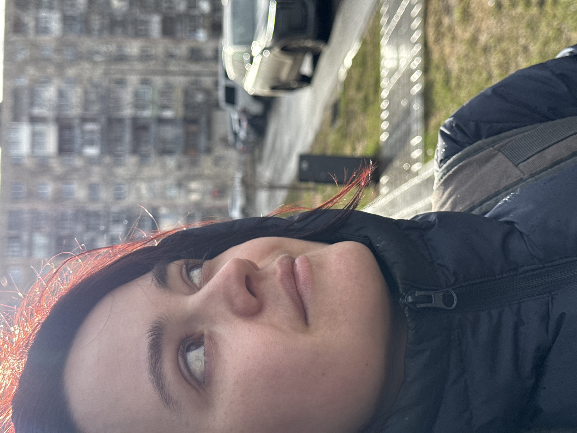 The image, oriented vertically, captures a close-up, eye-level portrait of a young woman outdoors under bright sunlight. Her face, in the foreground, is turned slightly to her right, and she is looking upwards. Her dark hair has strands around her face highlighted with a reddish glow from the sun. She wears a dark, possibly navy or black, puffy jacket, with a visible zipper closure on the front. A grey backpack strap is visible over her right shoulder.
In the blurred background to her left, a large multi-story building with numerous windows suggests an urban setting, possibly an apartment complex. To her right, a street or sidewalk with blurred parked cars is visible, along with a patch of green grass or dirt. Bright, circular bokeh effects are prominent in the background, indicating strong sunlight reflecting off surfaces, possibly a fence or foliage.
The time of day appears to be daytime, with clear, sunny weather. The woman's expression is calm and contemplative, as she gazes upwards. There is no visible text in the image. The scene is consistent with an urban environment in Izhevsk, Russia.