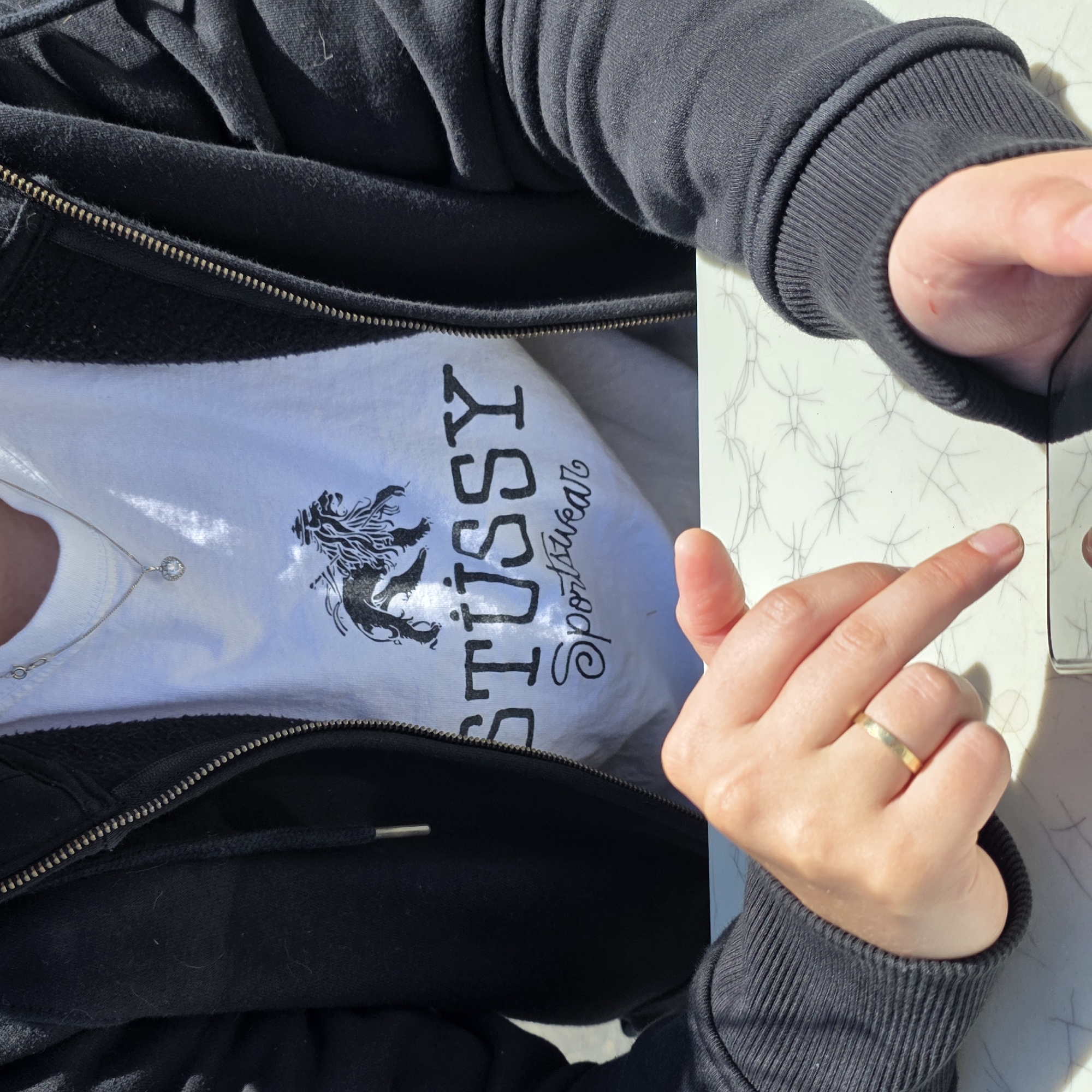 A person's torso and hands are visible, seated at a table with a patterned surface. They are wearing a black zip-up hoodie over a white t-shirt with a black graphic print that includes a lion and the words "STUSSY Sportswear". A silver necklace with a heart-shaped pendant is worn around their neck. The person is holding a smartphone and appears to be interacting with it, with one hand resting on the table and the other finger pointing downwards towards the device. A gold ring is visible on the ring finger of the left hand. The setting appears to be outdoors, with bright sunlight casting shadows. The exact location in Cannon Beach, USA is not specified.