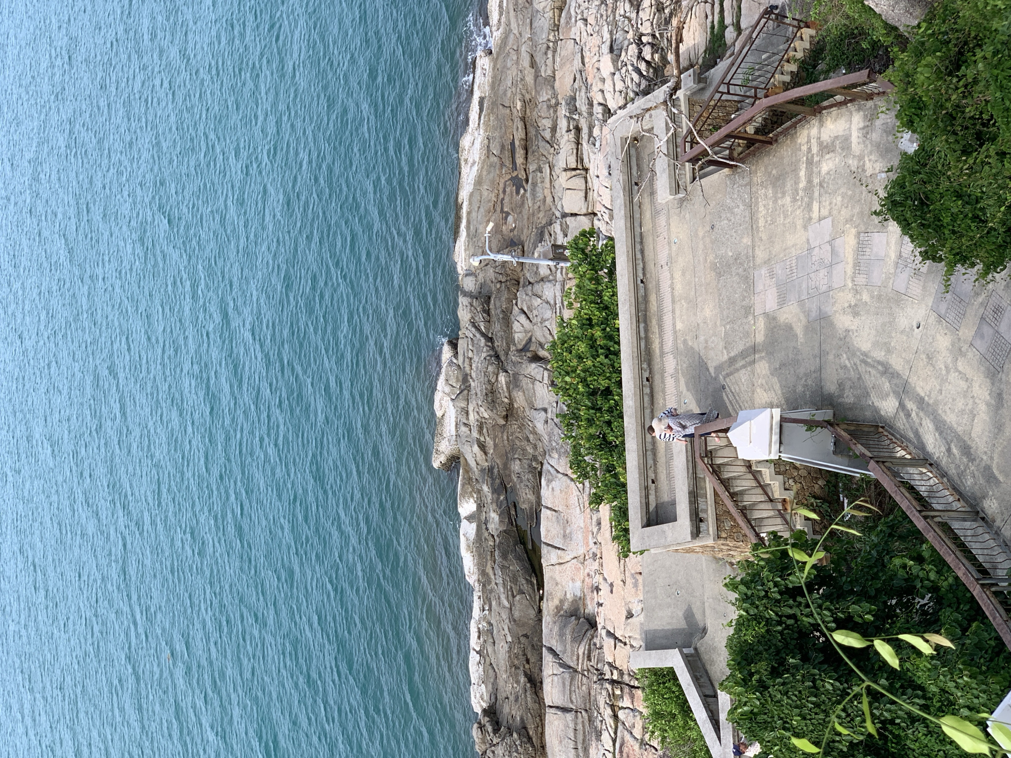 A person is visible on a concrete pathway next to a rocky coastline. The pathway has stairs and metal railings. The person appears to be looking out at the water. The scene depicts a serene coastal environment with lush green vegetation and calm blue water. The lighting suggests it is daytime. The location is Baan Chaweng Noi, Thailand. There are no visible text elements in the image.