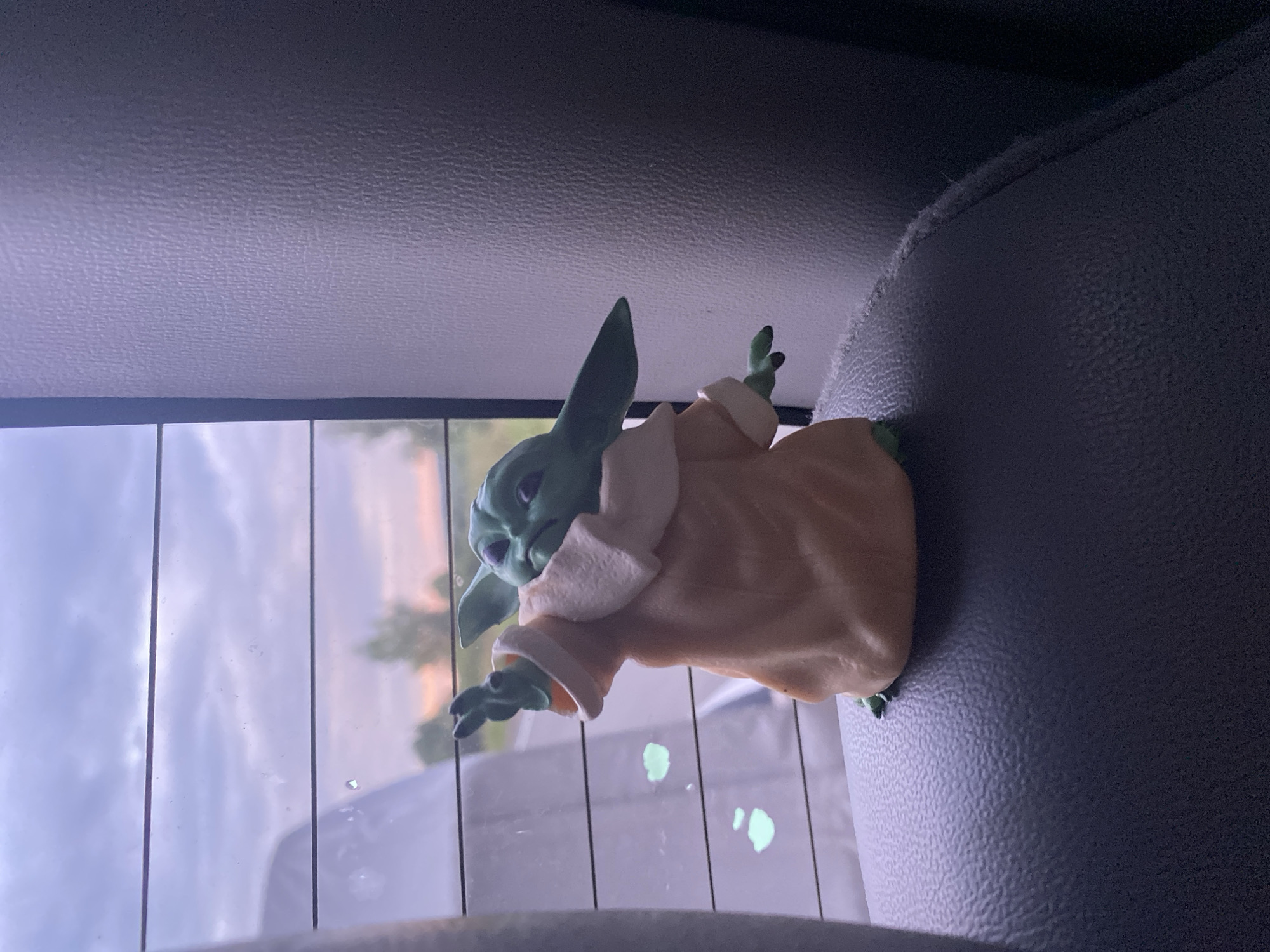 An action figure of Grogu, also known as Baby Yoda, is positioned on the textured dark gray headrest of a car seat. The figure is depicted in a light tan robe with a cream-colored collar. Its left arm is extended upwards and to the left, with its index finger pointing. The figure's head is turned to its right, with large ears and green skin.
Behind Grogu, a car window is visible, featuring horizontal defroster lines. Through the window, a soft, cloudy sky is visible, with the sky transitioning to soft pink and orange hues near the horizon, suggesting either sunrise or sunset. Sparse trees can be seen in the distance. There are two smudges of light green on the window, near the bottom left of the figure. The right side of the image is dominated by the textured dark gray upholstery of the car's interior. There is no discernible information about the location of Balakhi, Russia within the image.