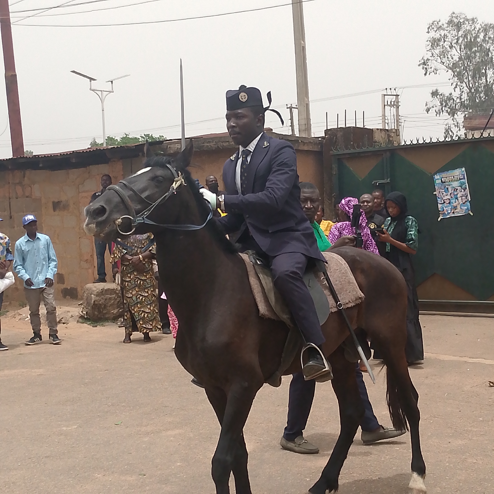 The image depicts a man in a dark blue suit and hat riding a dark brown horse down a paved street in Bukuru, Nigeria. The horse has a white blaze and is equipped with a bridle and saddle. The rider is wearing light-colored gloves and black shoes. A narrow, possibly ornamental sword is strapped behind the rider. Spectators are gathered on either side of the street, some taking pictures with their phones. Buildings and fences line the street. A street lamp and power lines are visible in the background. A poster is affixed to the green gate.