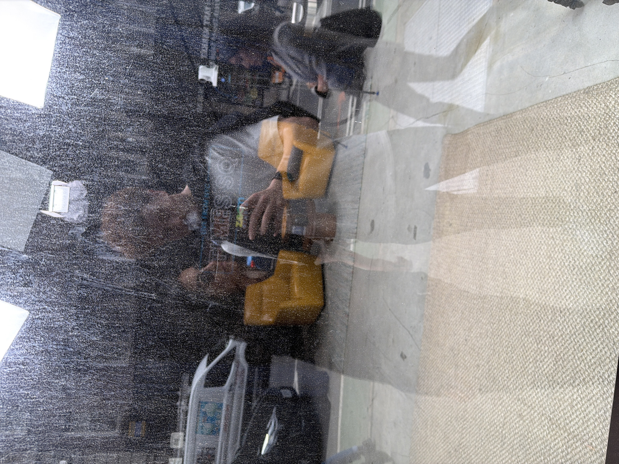This appears to be a photograph taken through a dirty or water-spattered window. A person is visible in the reflection, holding what seems to be a yellow utility container. Visible behind the person are camera equipment and a bottle of cleaning product. Outside the window, there's a view of a concrete floor and a textured rug. Specific location details within New York City cannot be determined from the image.