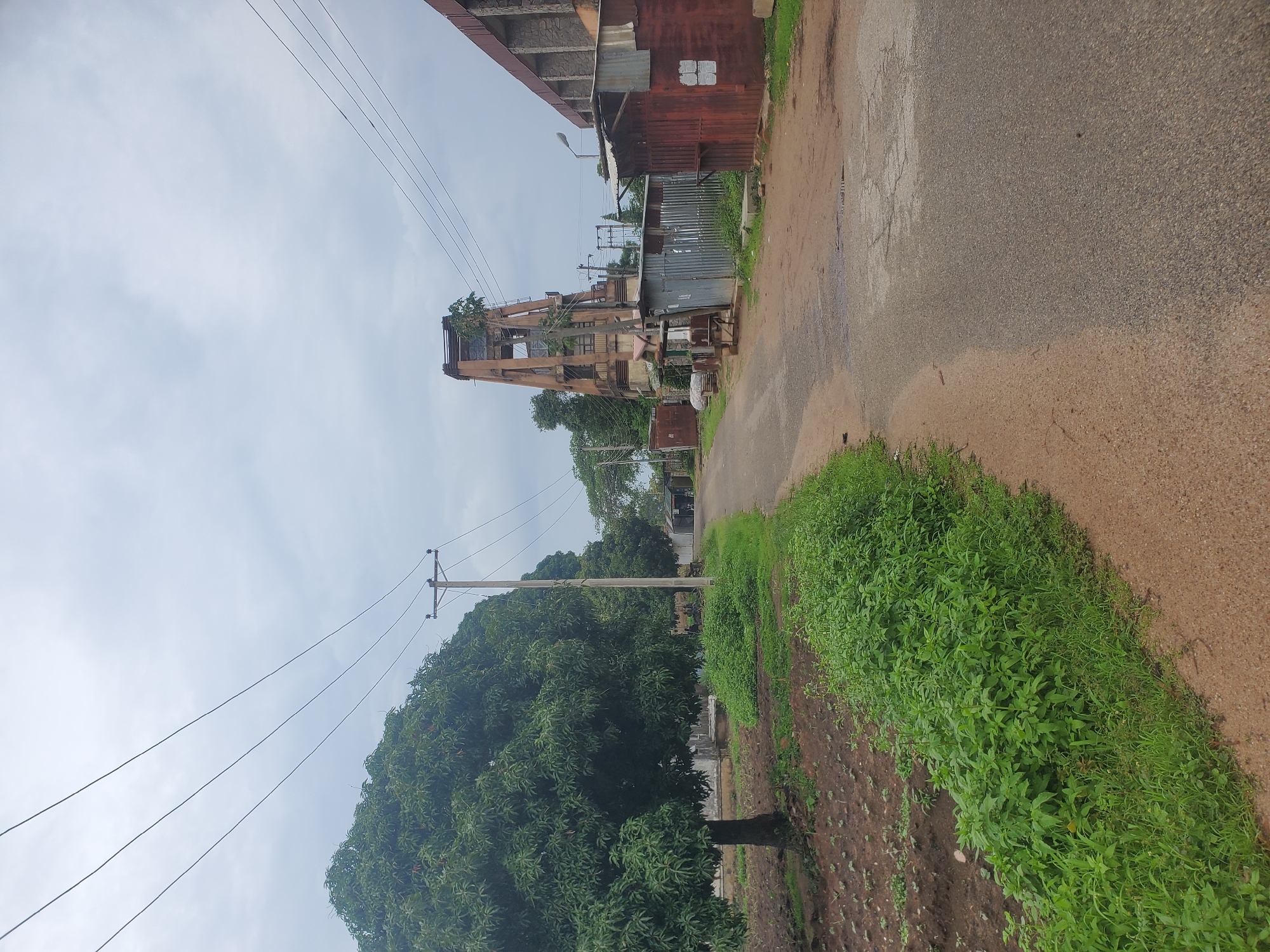 The image depicts a street scene in Jos, Nigeria, on an overcast day. On the left side of the paved road, there is a cultivated field with young plants, bordered by lush green vegetation and several large trees, one of which is a mature mango tree. A wooden utility pole with power lines runs alongside the field. Further down the road, on the right, is a distinctive tall, unfinished concrete structure resembling a tower. Adjacent to this structure are various makeshift buildings constructed from corrugated metal and wood, some appearing to be shops or stalls. One building on the far right has a more substantial, though unfinished, stone facade. The road itself is asphalt with patches of dirt and puddles of water. There are no people or animals visible in the image.