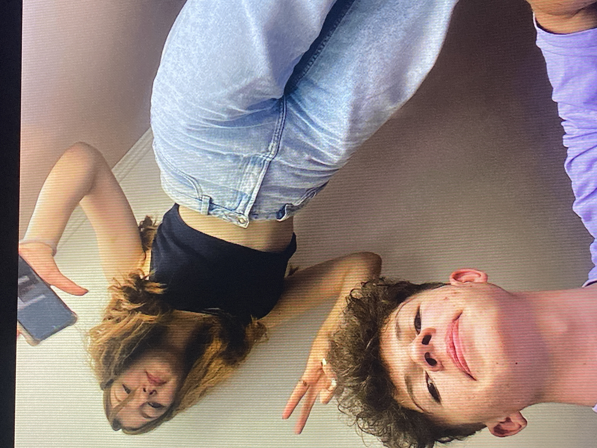 In a low-angle, indoor shot with a textured, cream-colored wall as the backdrop, two individuals are captured. The upper figure, a young woman with long, reddish-brown hair, is holding a smartphone, possibly taking a selfie. She is wearing a black crop top and light blue, wide-leg jeans. Her body is angled, and her face is partially visible, looking towards the phone.
Below her, a young man with curly brown hair is positioned closer to the camera. He is looking upwards and making a peace sign with his right hand, with two fingers extended. He appears to be wearing a purple t-shirt.
The image's orientation is unusual, suggesting it was taken from a high vantage point looking down, or that the subjects are positioned in a way that creates this perspective. There are no discernible landmarks or elements that would indicate the specific location within Poznan, Poland, or any other city. The scene is casual and appears to be a candid moment.