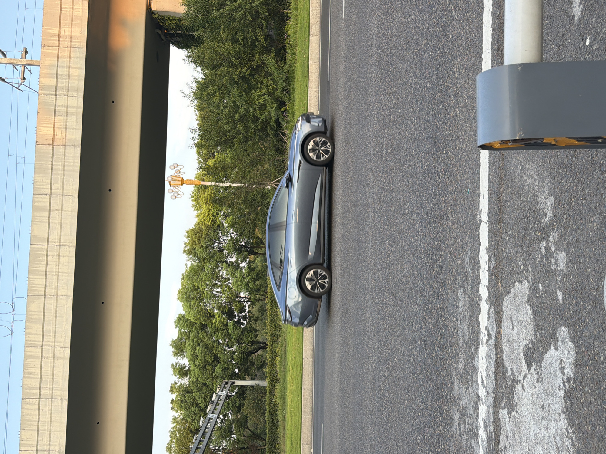 A grey sedan is driving on a paved road under a concrete overpass. The overpass has overhead wires and is supported by pillars. Trees line the side of the road behind a grassy median. The sky is clear and blue. A lamppost with multiple globes is visible on the left side of the road. The scene appears to be an urban or suburban area, possibly in Jiangjin, China, given the context.