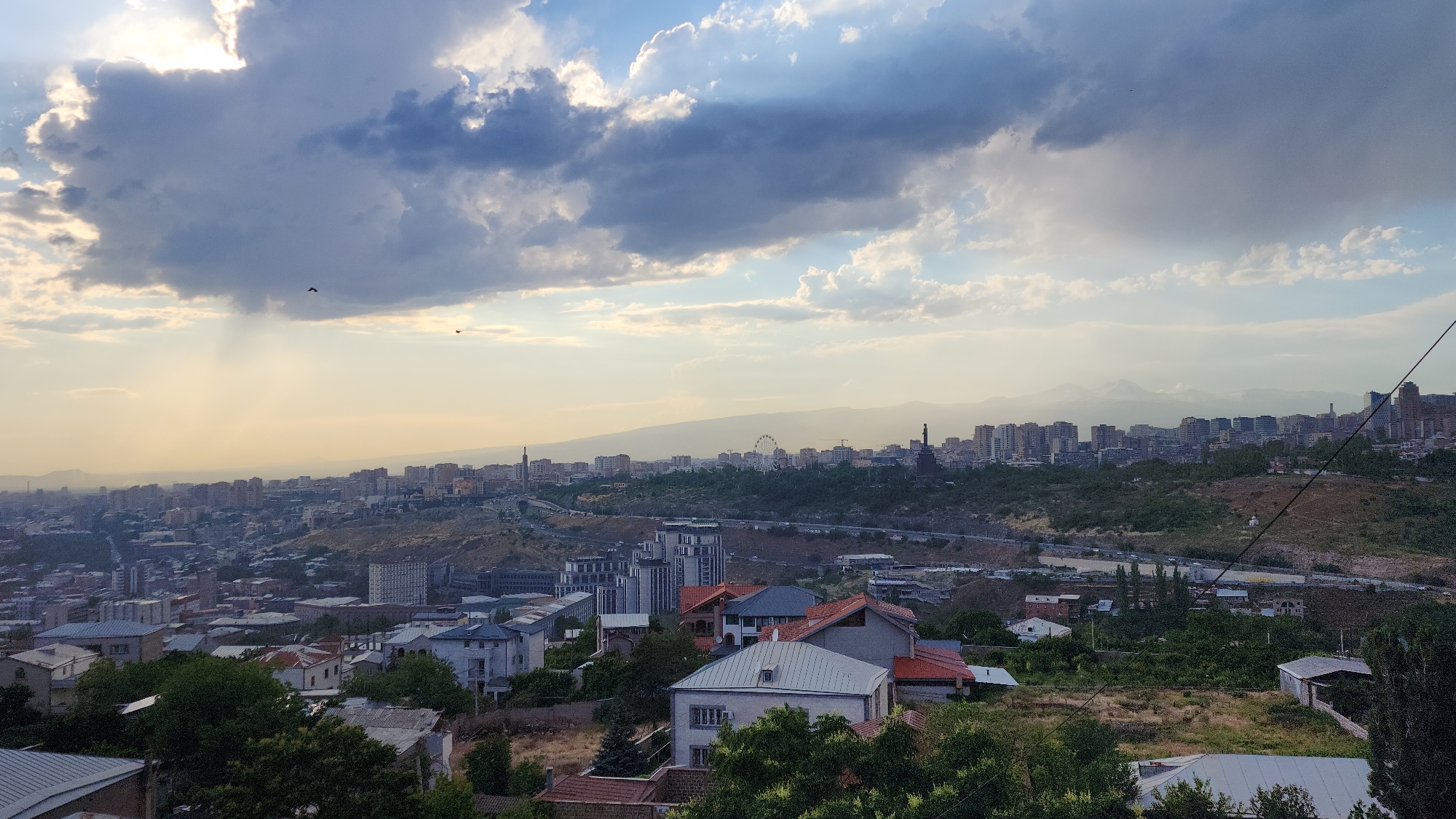 The image shows a panoramic view of Yerevan, Armenia, likely taken from a higher vantage point such as the Cascade complex or a nearby hill. In the foreground, several residential buildings with red and grey roofs are visible, nestled among lush green trees. The middle ground is dominated by a sprawling cityscape, characterized by apartment blocks and buildings of varying architectural styles, spread across undulating terrain. A road with vehicles can be observed winding through the city. In the distance, the silhouette of a Ferris wheel and a prominent statue, possibly the Mother Armenia monument, are discernible amidst the urban landscape. The background features a hazy mountain range, with snow-capped peaks visible in the far distance, indicating a clear day with a sense of depth. The sky is a dramatic mix of light and dark clouds, with shafts of sunlight breaking through, creating a dynamic and atmospheric effect. Two birds are in flight against the sky. The overall impression is one of a vast urban environment set against a natural backdrop, with a moody sky adding to the visual appeal of the scene.