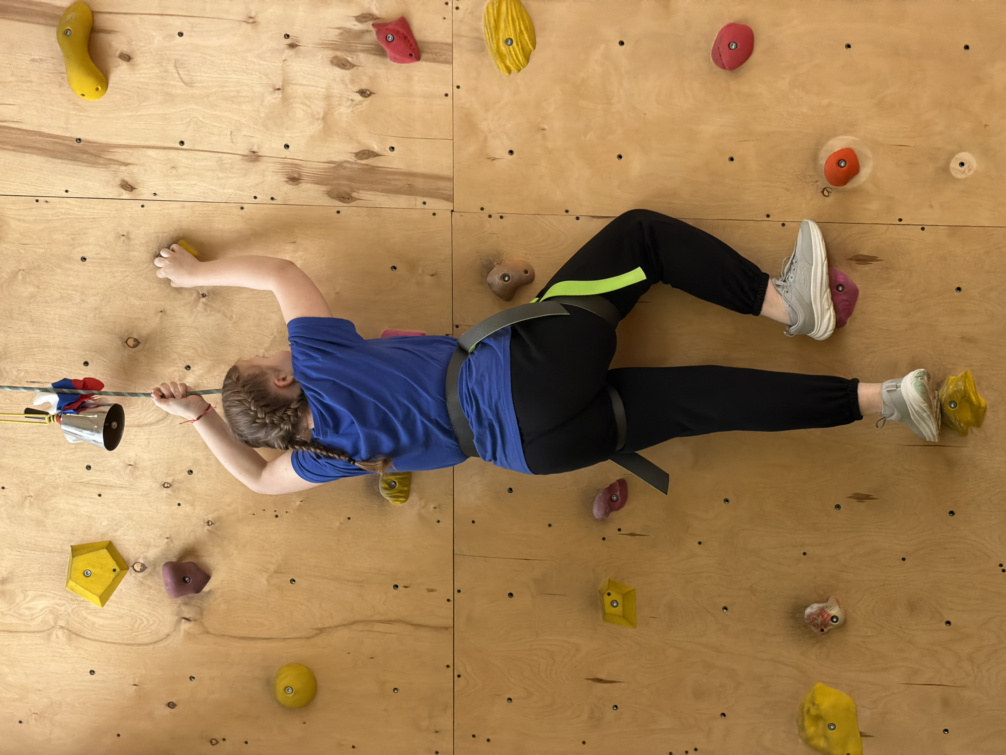 An individual is depicted actively climbing an indoor rock wall, likely situated in Izhevsk, Russia. The person, with braided hair, wears a blue t-shirt, black pants with a distinctive neon green stripe, and light grey sneakers. A black climbing harness is secured around their waist and thighs.
The climbing wall itself is constructed from light-colored wooden panels, fitted with a variety of brightly colored climbing holds in hues of yellow, red, orange, brown, and purple. The climber's left hand is gripping a rope, from which a silver bell, adorned with red, white, and blue ribbons, hangs – typically signifying the completion of a climbing route. Their right hand is positioned on a yellow climbing hold, and their feet are also placed on other holds, indicating a moment of ascent. The scene captures a focused athletic activity in a dedicated indoor climbing environment. There is no visible text in the image.