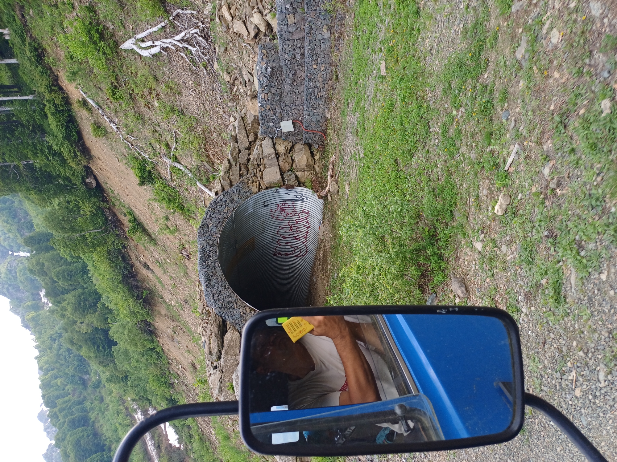 The media file is a photograph taken from the perspective of someone in a vehicle, looking towards a hillside. In the foreground, a blue side mirror of the vehicle reflects a man wearing a white t-shirt and holding a yellow note. The man appears to be looking down at the note. Beyond the mirror, a large corrugated metal pipe is visible, embedded in the hillside. The pipe appears to have red graffiti on its interior. The hillside is covered with green vegetation and rocks, with some dead trees and branches scattered across the slope. Further up the hill, there are more rocks and some structures made of stacked stones. The scene suggests an outdoor setting, likely in a rural or mountainous area, possibly under overcast skies given the diffused lighting. The presence of the corrugated pipe and the gabion walls suggests a landscape that has been modified for drainage or infrastructure. The overall impression is one of a rugged, natural environment, possibly a construction or maintenance area.