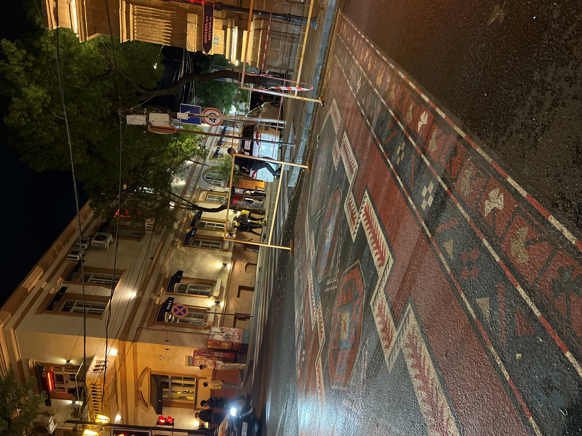 The image captures a street scene at night in Yerevan, Armenia. The street surface is wet, reflecting light from the street lamps and nearby buildings, and is painted with a large, patterned design resembling a traditional Armenian rug.
Buildings line the left side of the street, illuminated by warm lighting, with visible architectural details such as windows, air conditioning units, and signs. A traffic light is visible at the bottom of the frame, with red lights illuminated.
Several people are present, some standing near the curb and others crossing the street. Additionally, there is a street sign that says "Abovyan Str". A speed limit road sign with 40 on it is also visible.
