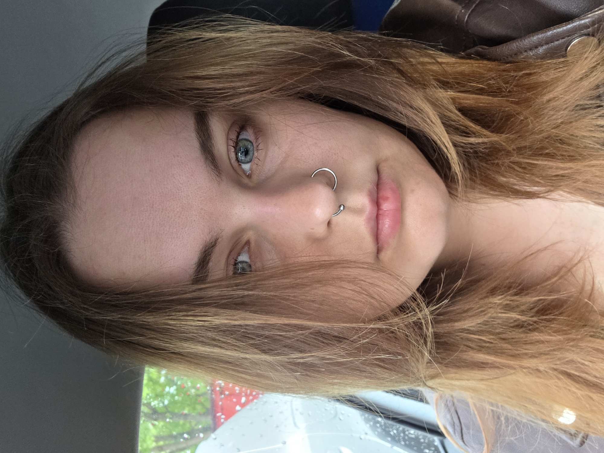 A young woman with light brown hair and blue eyes is looking directly at the camera. She has a silver nose ring. The background shows a car interior and a glimpse of a rainy day with trees and a building. The location is Unknown, Unknown.