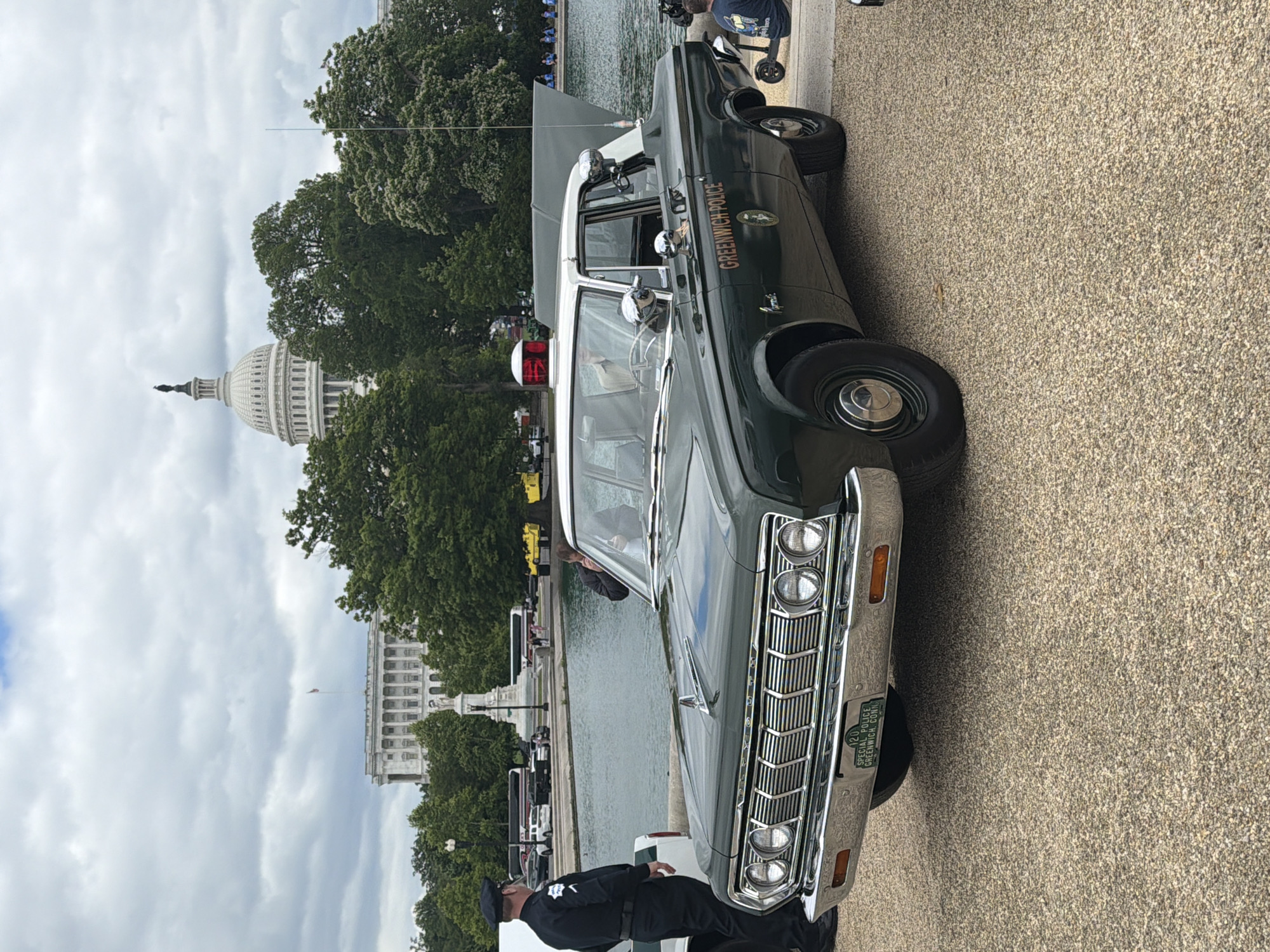 The image depicts a vintage dark green Greenwich Police car parked on a textured, possibly concrete, surface. A uniformed police officer is standing near the front of the vehicle. In the background, behind the car and police officer, there is a body of water with trees along the shoreline. In the distance, the United States Capitol Building is visible under a cloudy sky.