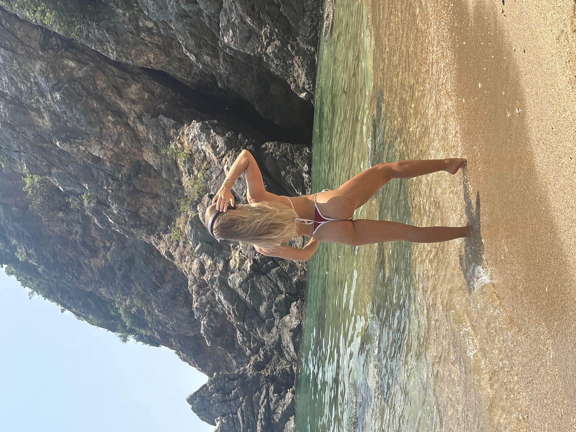A woman in a bikini stands on a beach with her back to the viewer. She is partially submerged in shallow, clear water. A rocky cliff face rises to her left. To her right is a sandy beach. The location cannot be identified as Đà Nẵng, Vietnam, without additional information.