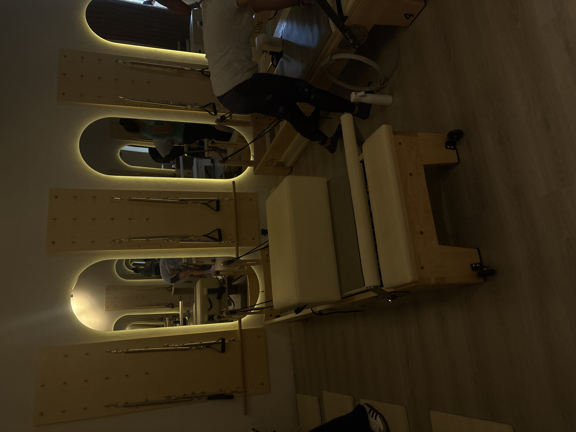 The image depicts the interior of a Pilates studio, likely located in Mexico City, Mexico. Several Pilates reformer machines are arranged in the room, each constructed from light-colored wood with beige upholstery. The machines are equipped with springs, ropes, and footrests, typical of this exercise apparatus. A person is shown seated on one of the reformers, indicating active use of the equipment. The studio features mirrors with rounded rectangular illuminated frames, mounted on the wall behind the machines, providing visual feedback for the users. The overall lighting is dim, creating a warm and relaxed atmosphere. The flooring appears to be made of wood or a wood-like material.