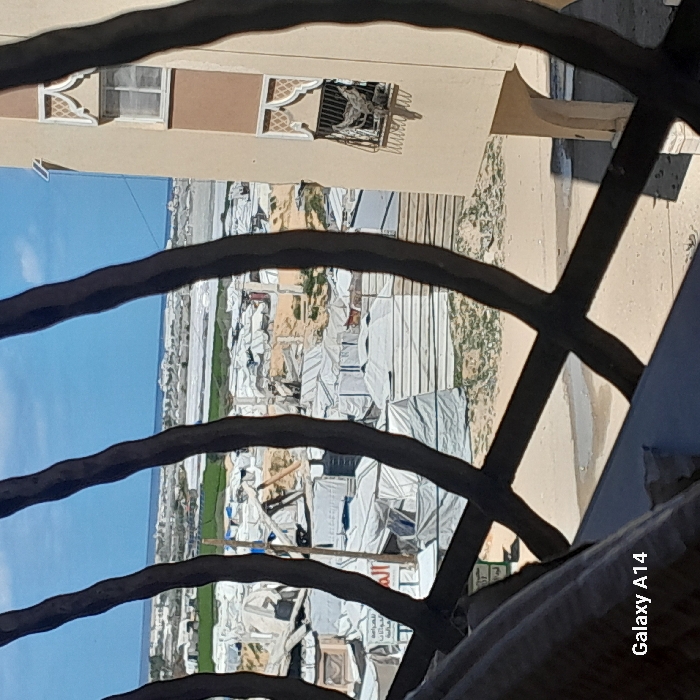 The image presents an outdoor scene in Khan Yunis, Palestinian Territory, viewed through dark, curved metal window bars. In the midground, a sprawling refugee camp occupies sandy ground, densely populated with numerous white and grey makeshift tents and shelters, some appearing partially collapsed. To the immediate right, a multi-story, light-beige building with distinctive arched window frames features a metal grate on an upper window from which laundry is hanging. The background displays a clear blue sky above the Mediterranean Sea, with a distant coastline. The scene is illuminated by bright daylight, casting visible shadows. The bottom left corner includes a "Galaxy A14" watermark. No individuals are discernible within the frame.
