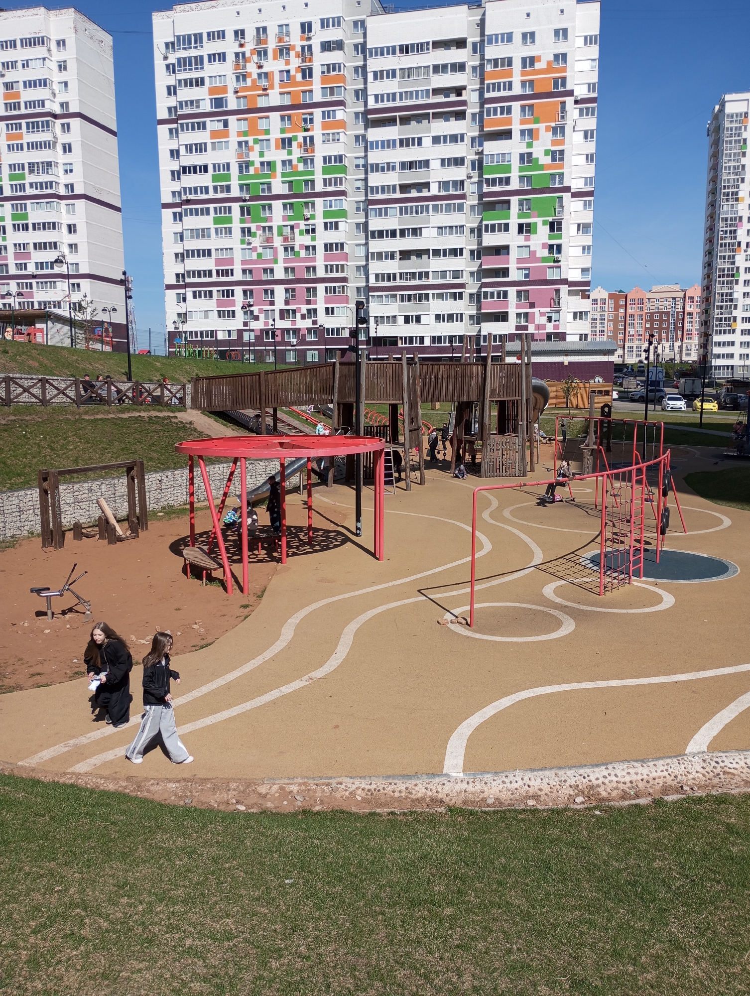 The image shows a playground in Izhevsk, Russia. Several high-rise apartment buildings are in the background, with colorful facades. The playground has various structures, including climbing frames, a red gazebo-like structure with benches, and other play equipment. Children are playing on the equipment. Two girls walk along the edge of the playground in the foreground. The ground covering is a mix of sand, tan rubber surfaces, and grassy areas. The day is sunny and the sky is clear. Several vehicles are visible in the distance beyond the playground and buildings.