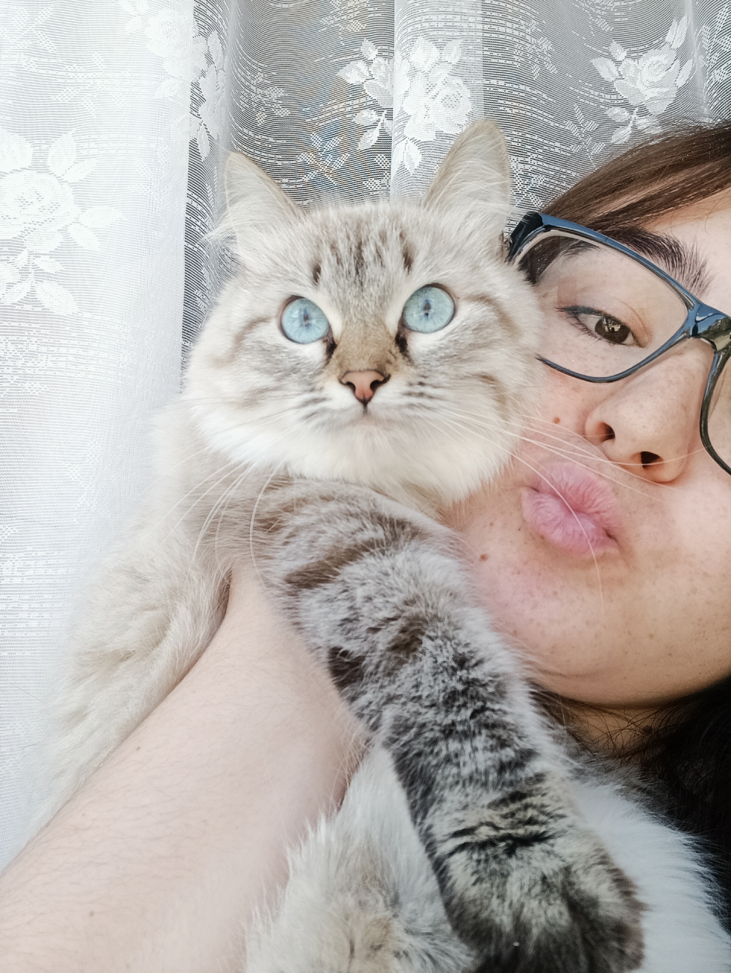 A selfie depicts a woman with brown hair and glasses holding a cat with striking blue eyes. The cat is positioned close to the woman's face. The background features a white curtain. It is not possible to determine the location as Noviy, Russia from the visual information provided.