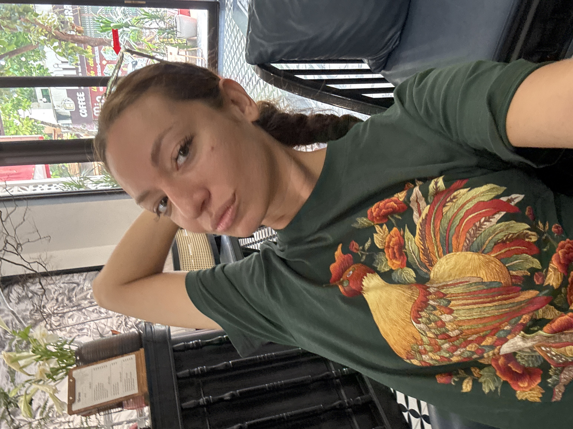 The image is a selfie taken by a woman indoors, likely in a café or restaurant in Phố Ràng, Vietnam, during the daytime. The woman, who has brown hair tied back in a braid, looks directly at the camera with a neutral expression. She is wearing a dark green short-sleeved t-shirt featuring a prominent and colorful design of a rooster surrounded by vibrant red and orange flowers. The rooster has a yellow body, a red comb and wattle, and multi-colored tail feathers in shades of red, orange, green, and gold.
The setting appears to be a cozy establishment. Behind the woman, a large window reveals an outdoor scene with lush green foliage, trees, and other buildings. A sign with the word "COFFEE" is clearly visible through the window, along with additional text that appears to be in Vietnamese, partially reading "o - cac ban."
To the woman's left, a wooden menu board resembling a clipboard is displayed near a vase holding white flowers, possibly lilies, and decorative dried branches. Dark, possibly rattan or wooden, furniture, including a dark blue upholstered cushion, is visible in the background. The floor features a distinctive black and white tiled pattern. The overall ambiance suggests a relaxed and casual environment.