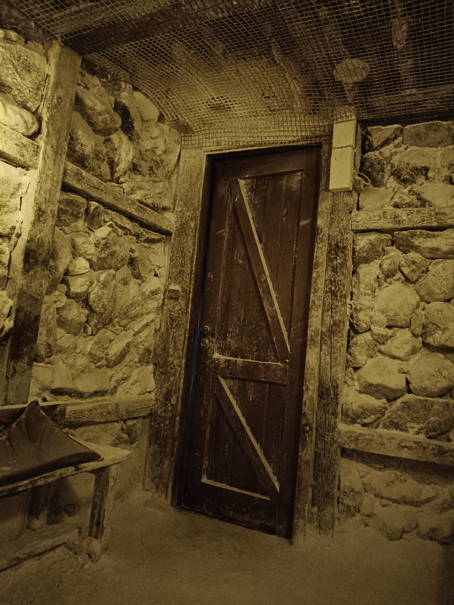 The image depicts the interior of a rustic, underground space, likely a cellar or a similar structure, with walls constructed from rough, unhewn stones. A dark wooden door with a diagonal plank design stands centrally in the frame, appearing weathered and aged. Above, a wire mesh ceiling is visible, suggesting a protective covering or ventilation system. The overall lighting is dim and sepia-toned, creating a somber atmosphere. The floor appears to be covered in dust or fine debris. To the left, a portion of a wooden bench with a metal scoop is visible, hinting at past use for storage or labor. This scene is located within the Museum of Aviation in Plovdiv, Bulgaria, specifically within the historical exhibit dedicated to the underground tunnels and shelters used during wartime.