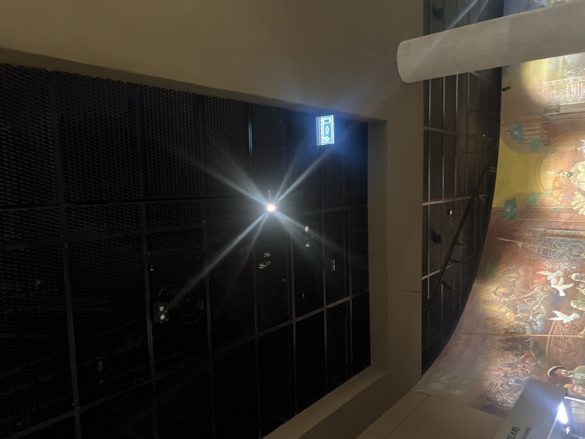 A bright light shines from the ceiling in the center of a large room, casting a starburst effect. To the right of the light, a green illuminated exit sign with arrows pointing both left and right is visible. The ceiling is a dark, grated structure with a grid pattern. Below the ceiling, a large, colorful mural depicting a historical battle scene covers a curved wall. The mural shows numerous figures in military attire, some on horseback, with banners and weapons. A tall, light-colored cylindrical pillar stands on the right side of the frame, obscuring part of the mural. A sliver of text is visible on the left side of the mural. The environment appears to be an indoor public space, possibly a museum or exhibition hall.