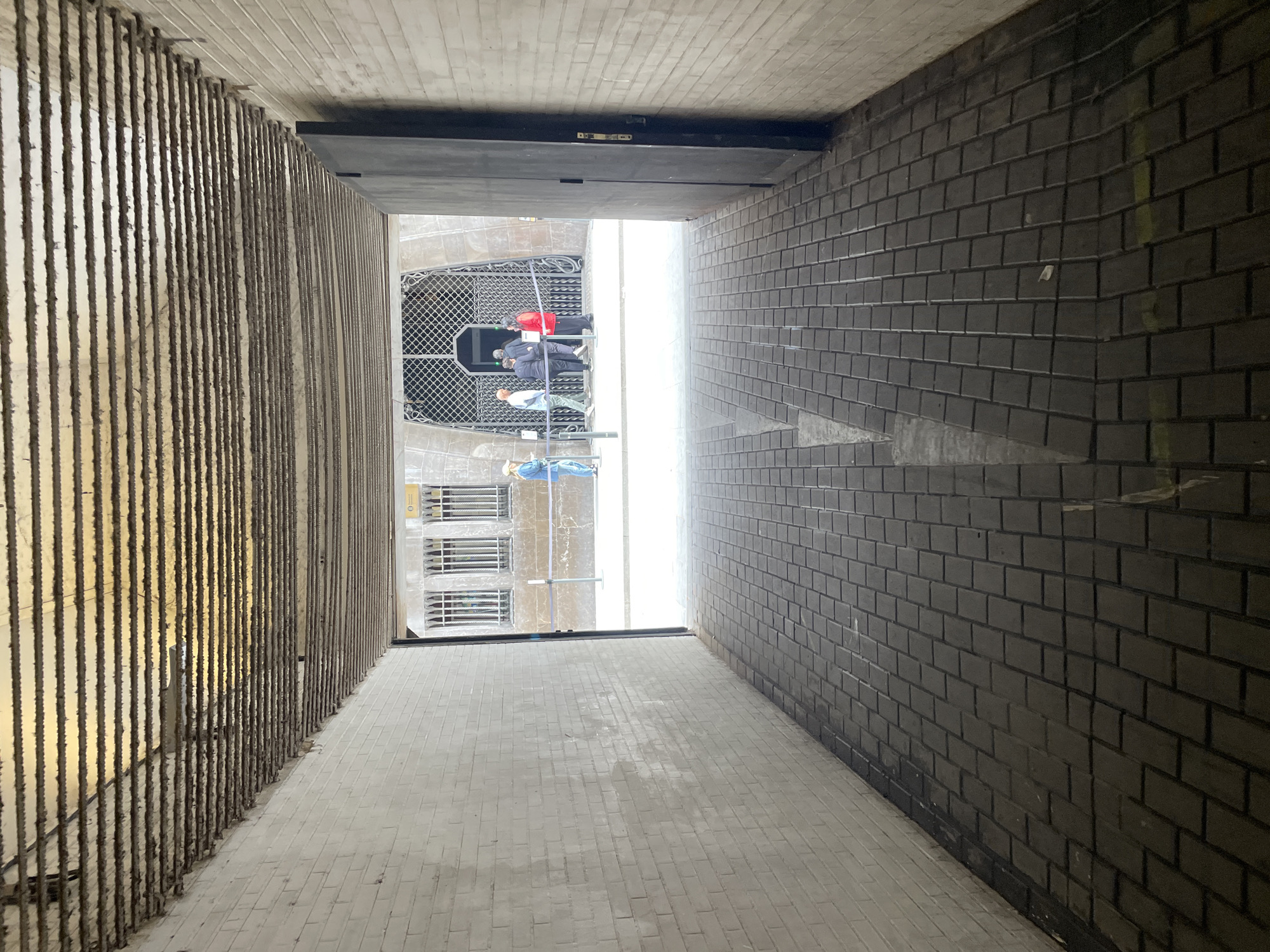 The image presents a view down a narrow alleyway in Barcelona, Spain. The left wall is constructed of vertical, textured concrete elements. The right wall is composed of dark gray brick. The floor is paved with light gray rectangular tiles. At the end of the alleyway, visible through an opening, are scaffolding, a metal grate, and several people on the scaffolding, one of whom is wearing a red cap. A building with windows is also visible. A dark horizontal barrier is across the top of the opening at the end of the alleyway. The perspective suggests the image was taken from above.