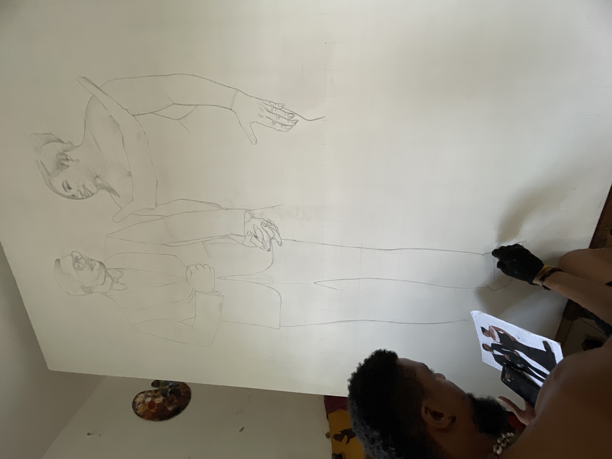 An artist is drawing a portrait on a large canvas using a smartphone as a reference. The drawing depicts a man in a suit standing, and a woman in the foreground reaching her hand. The scene is indoors with a painter's palette on the floor.