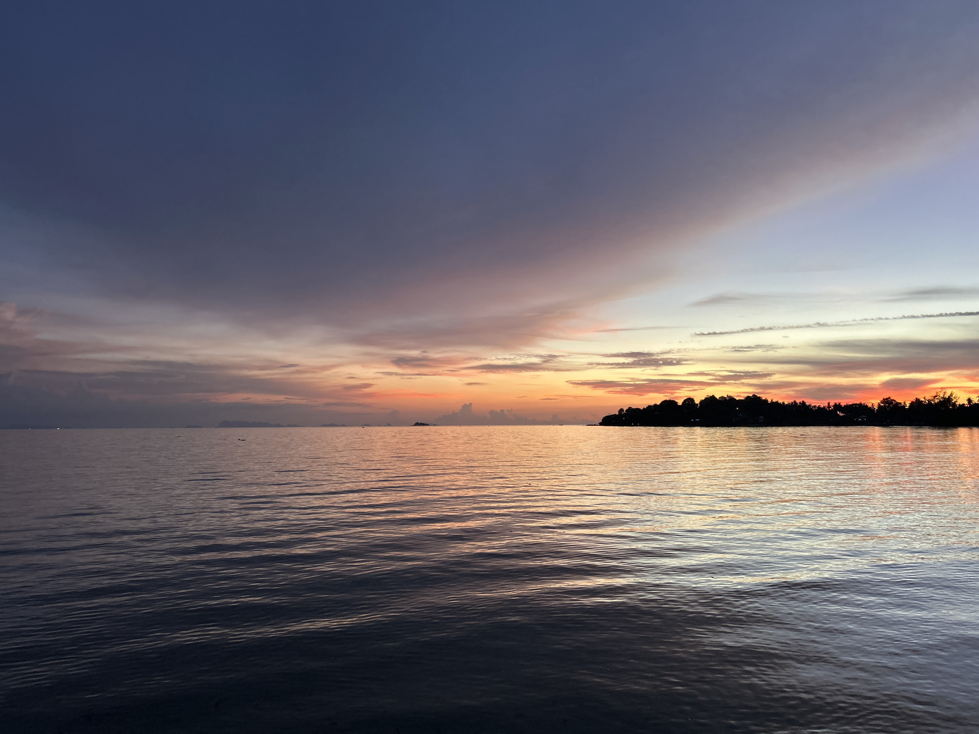 This is a landscape photograph taken at dusk in the Ko Pha-ngan District, Thailand. The image depicts a calm sea with gentle ripples reflecting the twilight sky. The sky displays a gradient of colors, transitioning from deep blue at the top to shades of purple, pink, and orange near the horizon. Silhouetted against the vibrant sky is a distant treeline, suggesting land or an island. There are no people or identifiable objects in the foreground, emphasizing the natural beauty of the seascape and the sky. The overall scene evokes a sense of tranquility and the peaceful transition from day to night.