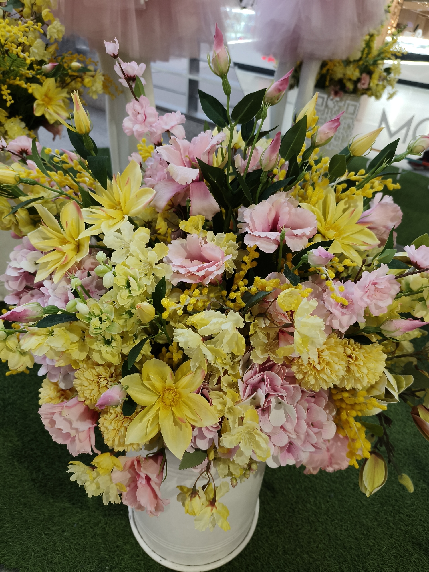 A vibrant, large bouquet of artificial flowers, primarily in shades of yellow and pink, is prominently displayed in a white cylindrical vase or bucket, set against a green artificial turf surface. The bouquet features a rich variety of floral types.
Dominant yellow flowers include large, open-faced blooms resembling dahlias or gerberas, dense clusters of chrysanthemum-like flowers, and numerous small, fluffy spherical flowers characteristic of mimosa, spread throughout the arrangement. Slender yellow buds are also visible. The pink flowers consist of multi-petaled, ruffled blooms akin to carnations or lisianthus, and smaller, clustered flowers that could be cherry blossoms or hydrangeas. Several delicate pink flower buds complement the arrangement. Green leaves are interspersed among the blooms, providing contrast.
The background is softly blurred but reveals elements of an indoor setting, likely a retail space or an event in Dnipro, Ukraine. Hints of white display structures or columns are visible, along with a piece of light pink tulle fabric, possibly part of a decoration or garment on display. Partial text, "MO", can be seen on a white surface in the mid-background. The overall impression is one of bright, cheerful decoration, evoking feelings of spring or celebration. No specific human activity is depicted; the focus is solely on the floral arrangement as a static display.