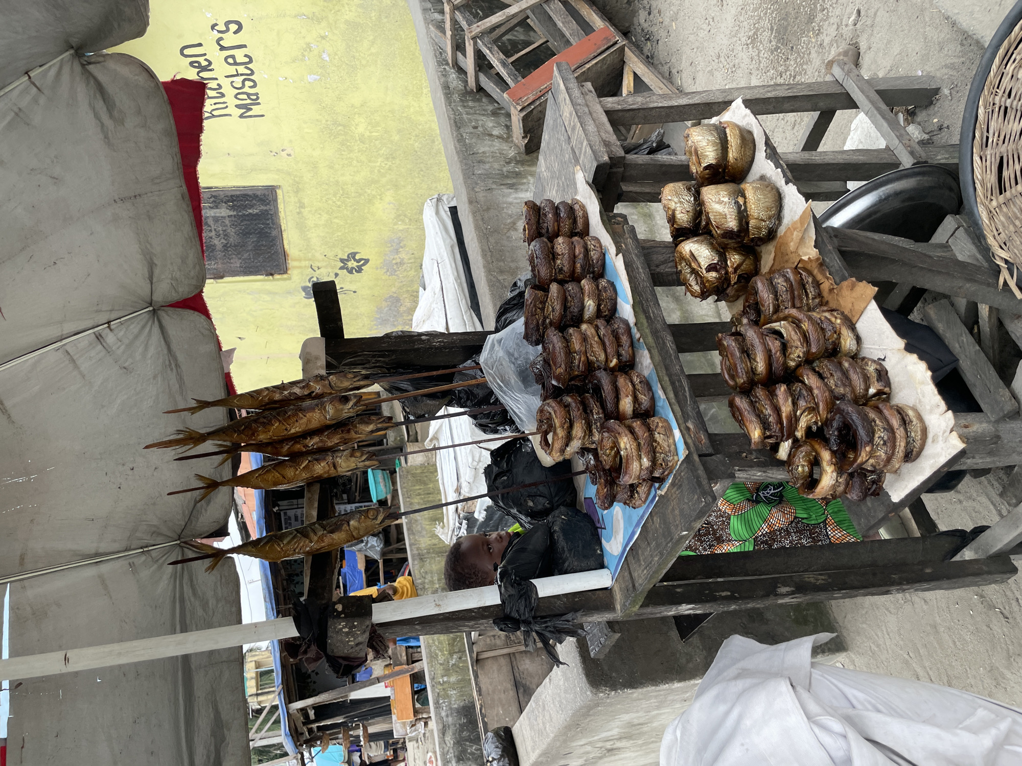 A street vendor in Ado, Nigeria, displays smoked fish for sale. The vendor's stall features a wooden table laden with various types of smoked fish. Hanging from sticks above the table are whole, elongated smoked fish. Stacked on the table are circular, ring-shaped smoked fish. A young child is visible sitting amongst the merchandise, wrapped in black plastic. The stall is partially covered by a large, light-colored umbrella, and a yellow wall with faded text reading "Kitchen Masters" is visible in the background. The ground is paved with sand or dirt. Wooden crates and other market paraphernalia are scattered around the scene.